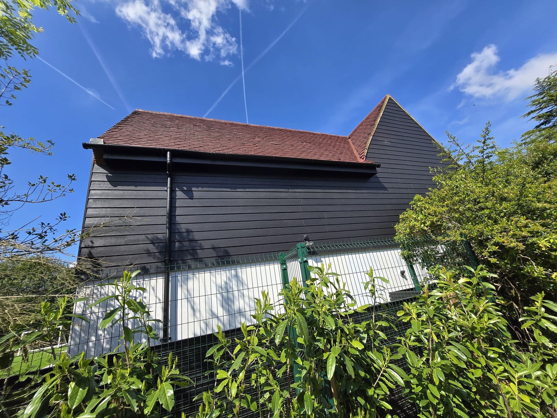 Dark barn-like building with a red tiled roof, against a blue sky with clouds and trees.