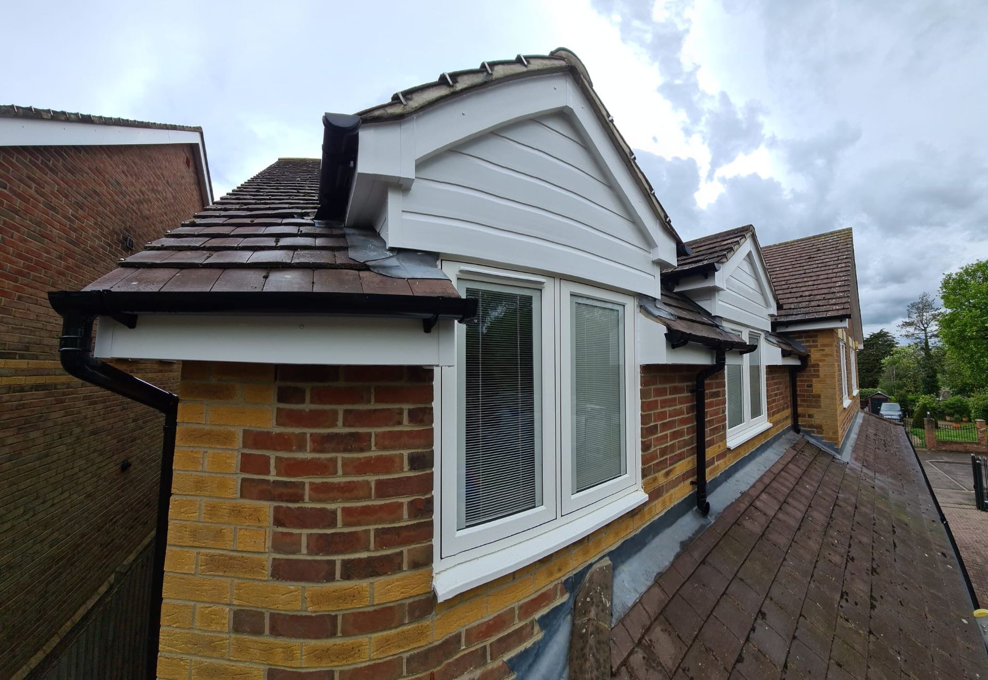 Exterior view of a house roof featuring brick walls, dark brown tiled roofing, and two white dormer windows with trim.