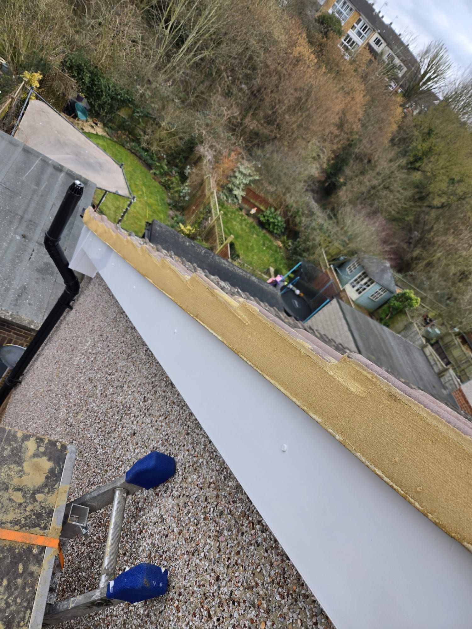 A high-angle view of a roof edge under repair, showing exposed wood framing next to a gray gutter above a grassy backyard.