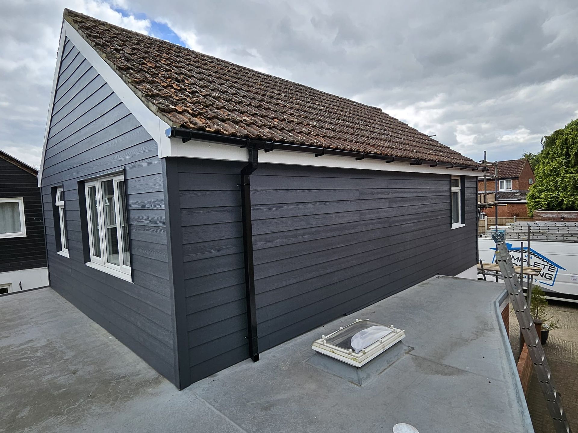 A gray-painted wooden outbuilding with a tiled roof, white window frames, and a flat-roofed section with a skylight.