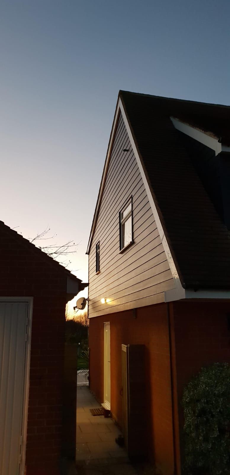 Narrow pathway between a brick building and a house with white horizontal siding and a dark roof at sunset.