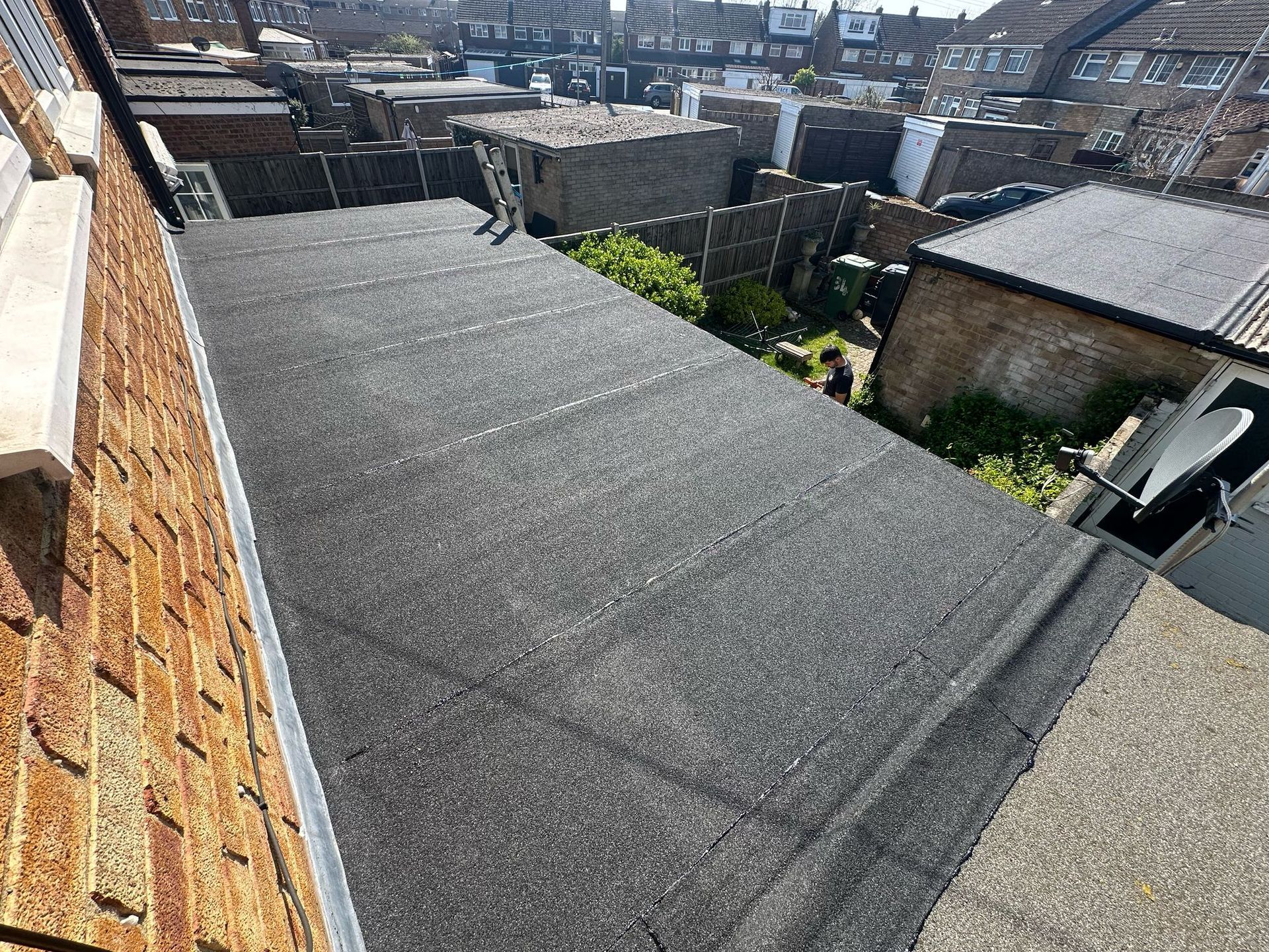 An overhead view of a newly installed, dark gray flat roof with visible seams, adjacent to a brick wall.