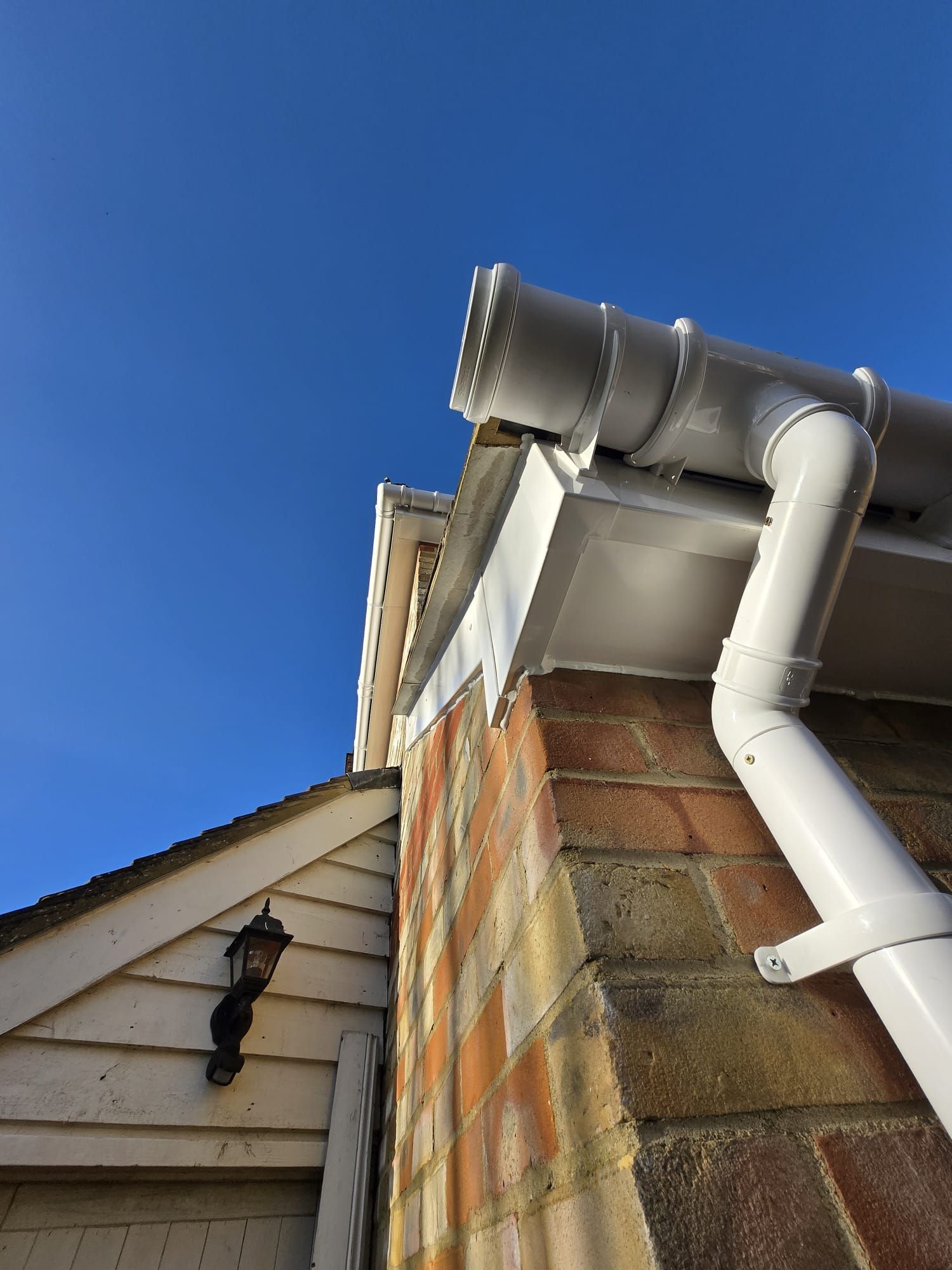Low-angle view of a corner of a brick building with white guttering, a downpipe, and a wall lamp against a blue sky.