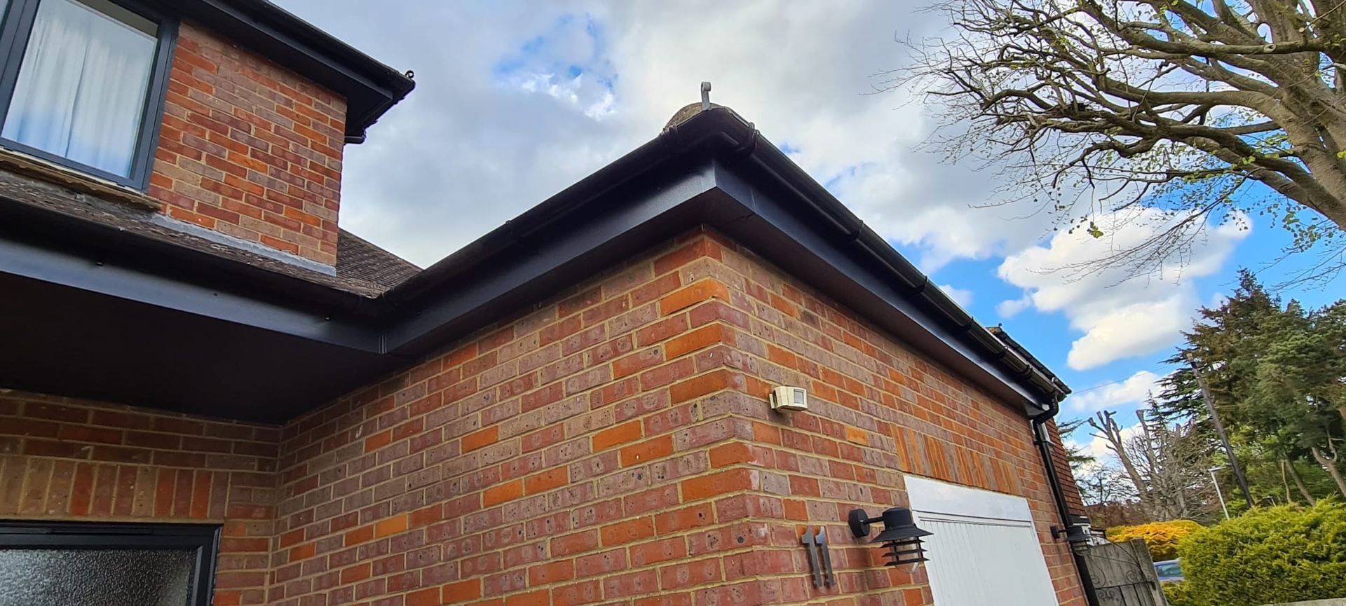 A brick house exterior with a dark roofline, soffits, and gutters under a partly cloudy sky.