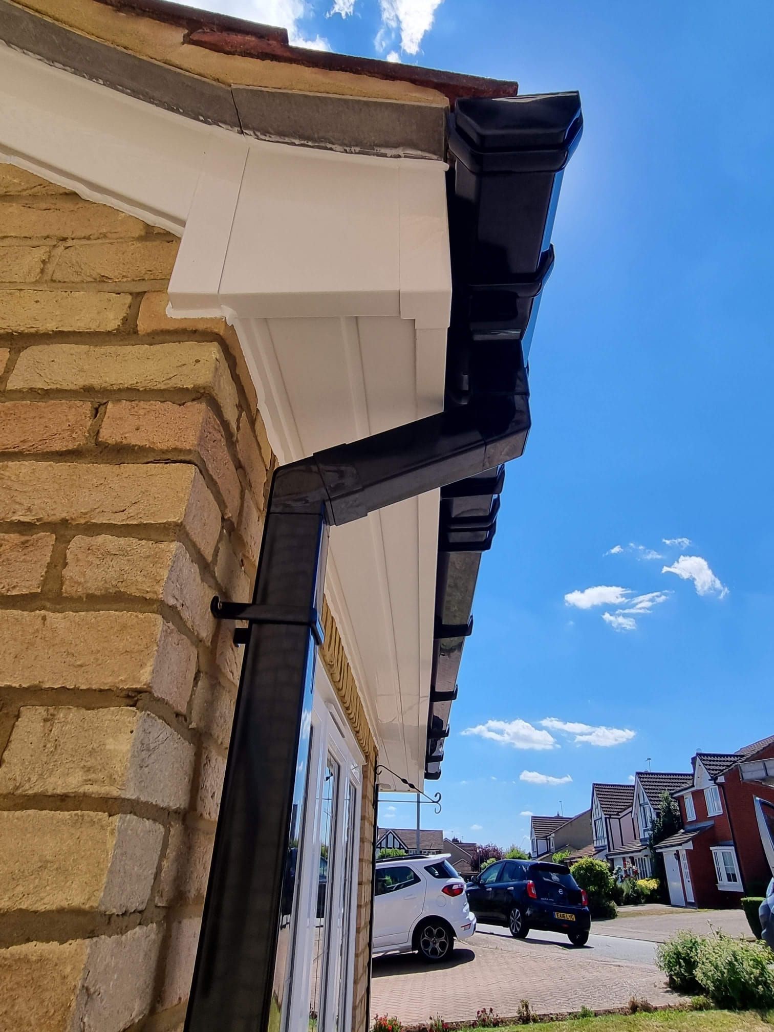 Black gutters and white soffit on a brick building against a blue sky with visible houses and cars.