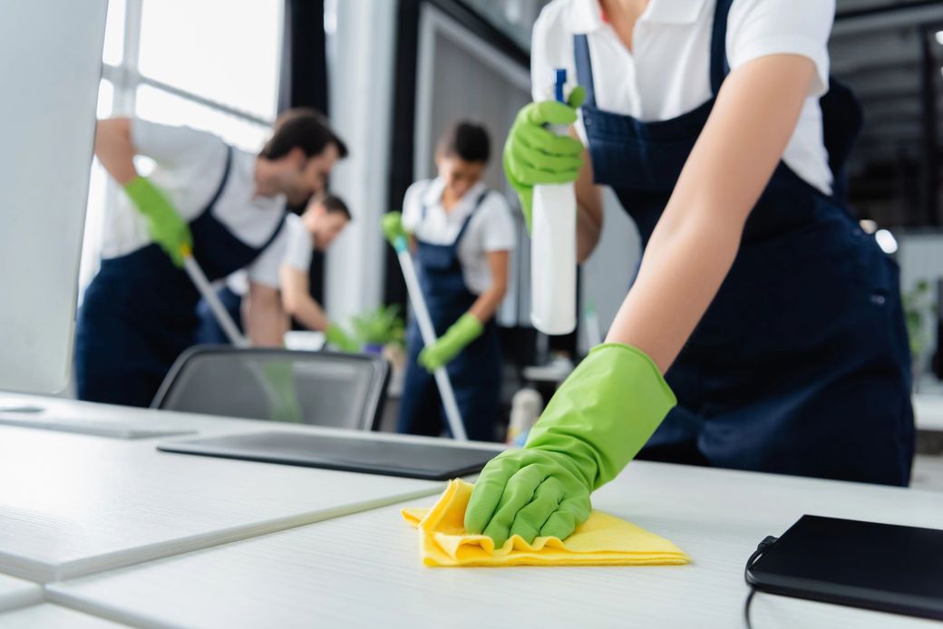 Persone che puliscono un tavolo con guanti verdi e panni gialli in un ufficio o in una mensa.