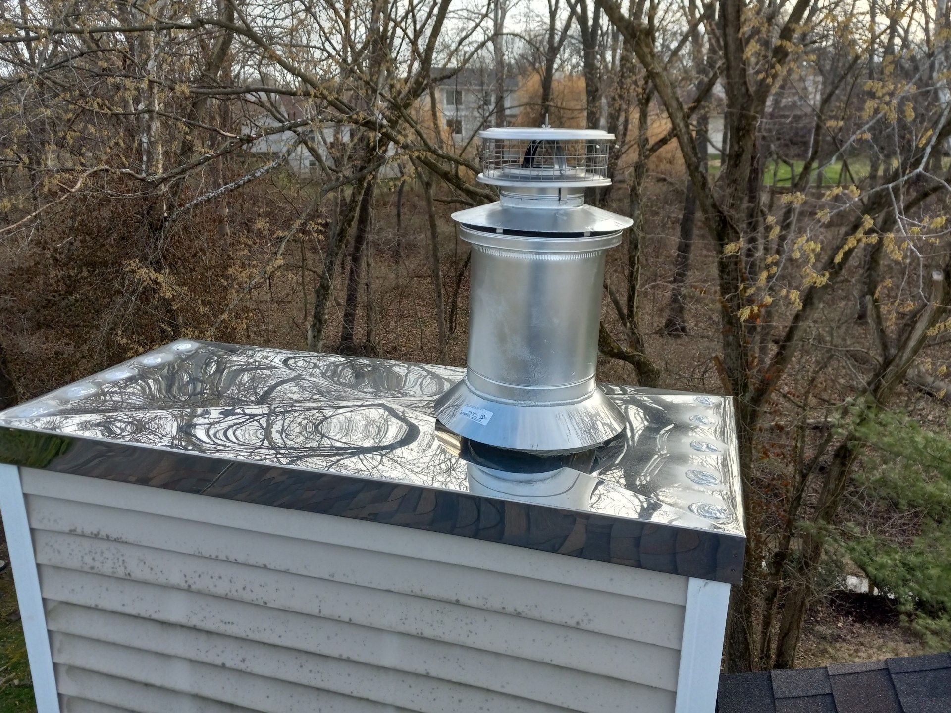 Silver chimney vent on a white-sided roof with bare trees in the background