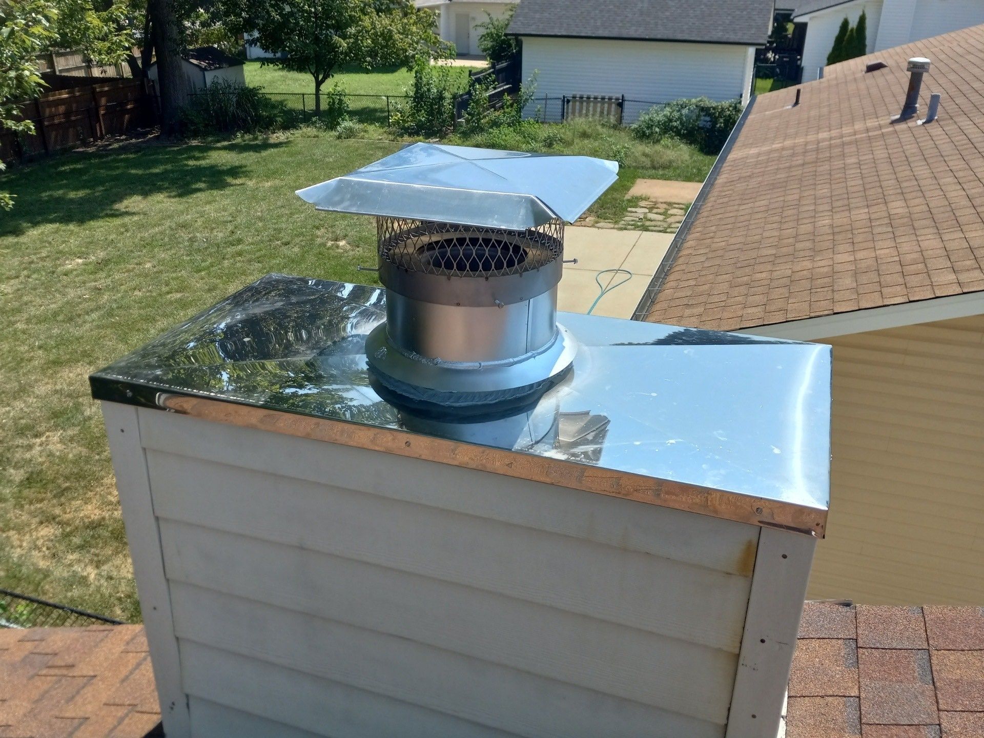 Rooftop chimney vent with shiny metal cap on a sloped shingle roof, backyard and neighboring house in background