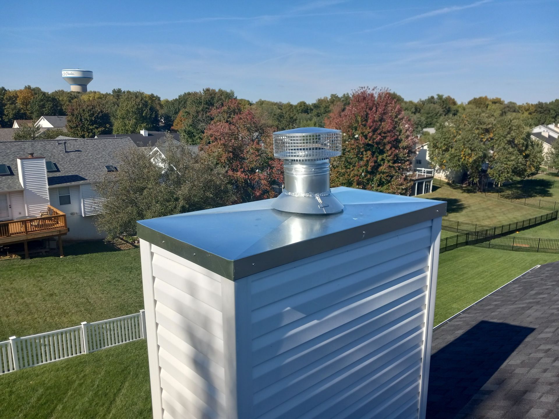 Rooftop vent pipe on a white building with a black roof, overlooking houses and trees under a blue sky