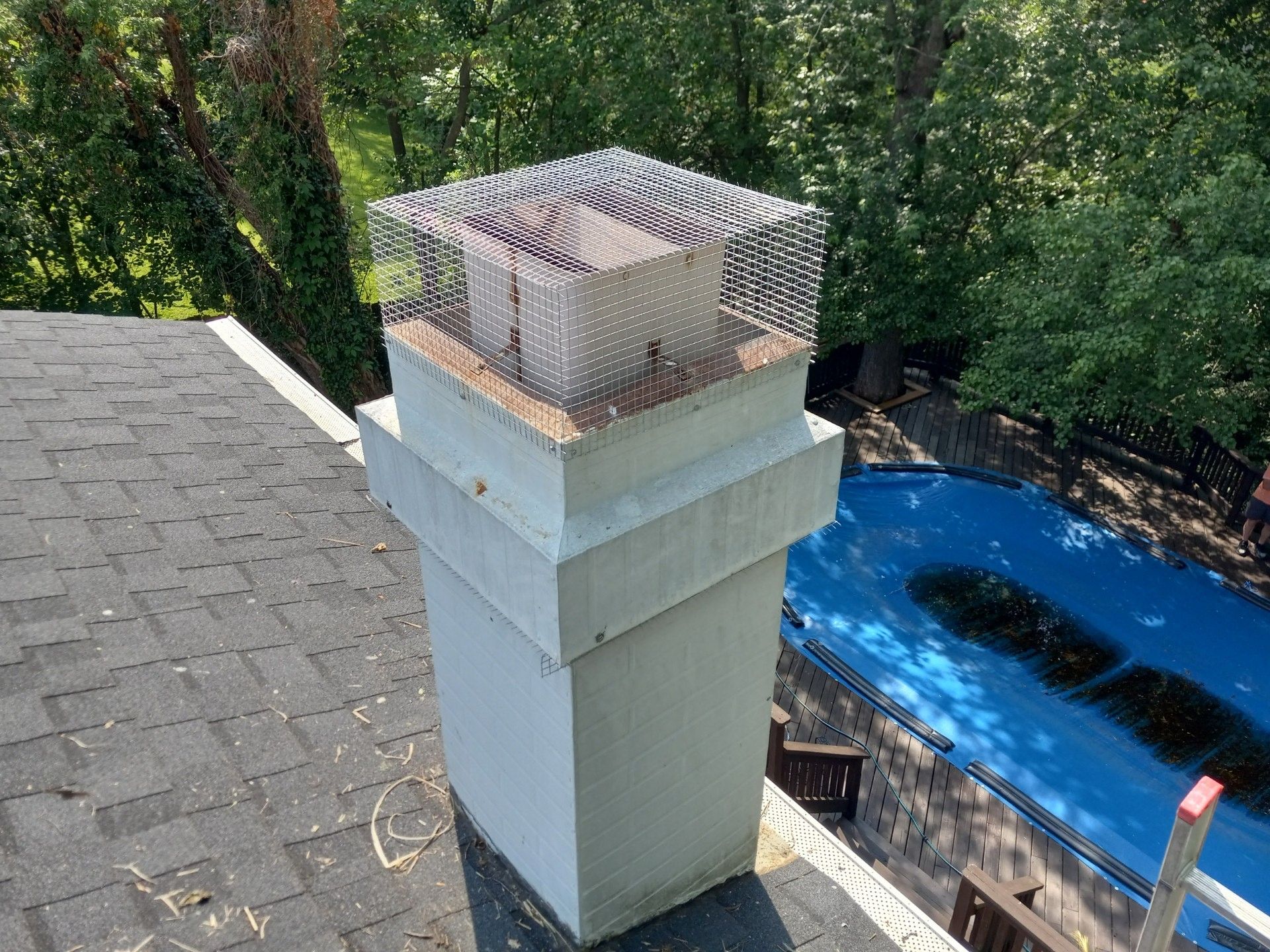 Backyard chimney with a metal cap on a shingled roof, beside a blue pool and trees