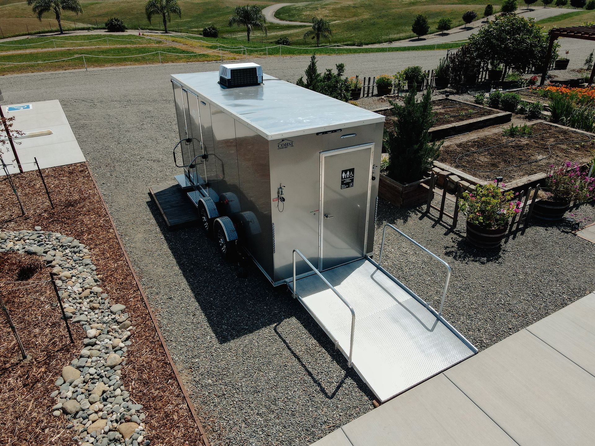 Portable white utility trailer with a ramp, parked on gravel in a landscaped outdoor area.