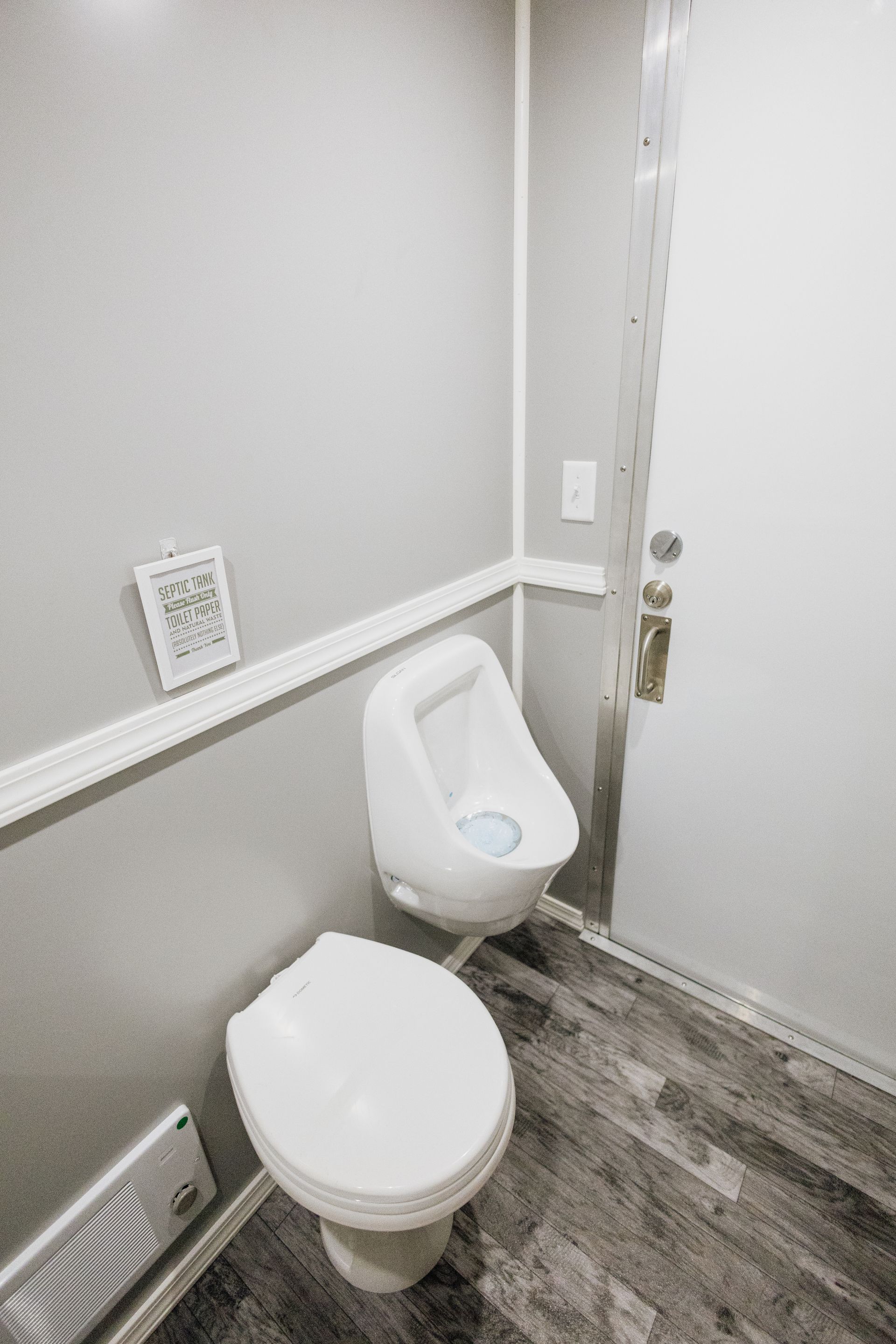 Small restroom with two white toilets and gray wood-look flooring, viewed toward a closed door