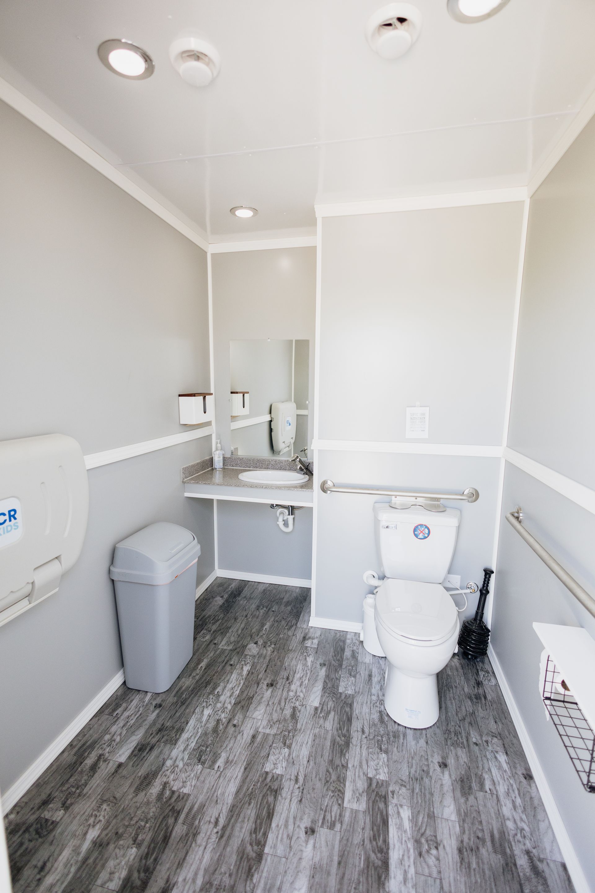 Compact white bathroom with toilet, sink, mirror, trash can, and gray wood-look floor
