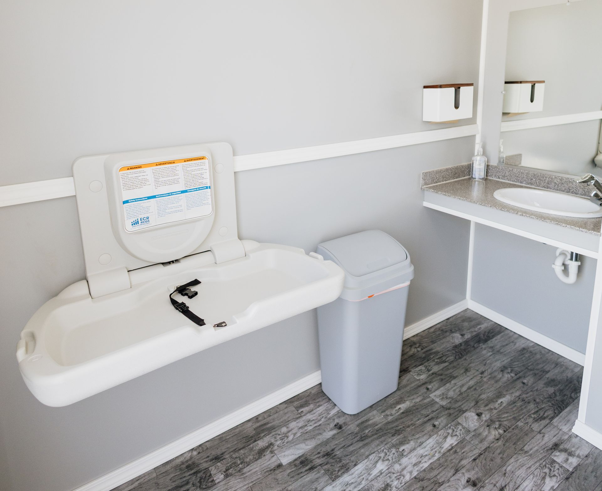 Accessible bathroom with wall-mounted sink, grab bar toilet seat, trash bin, and wood-look floor