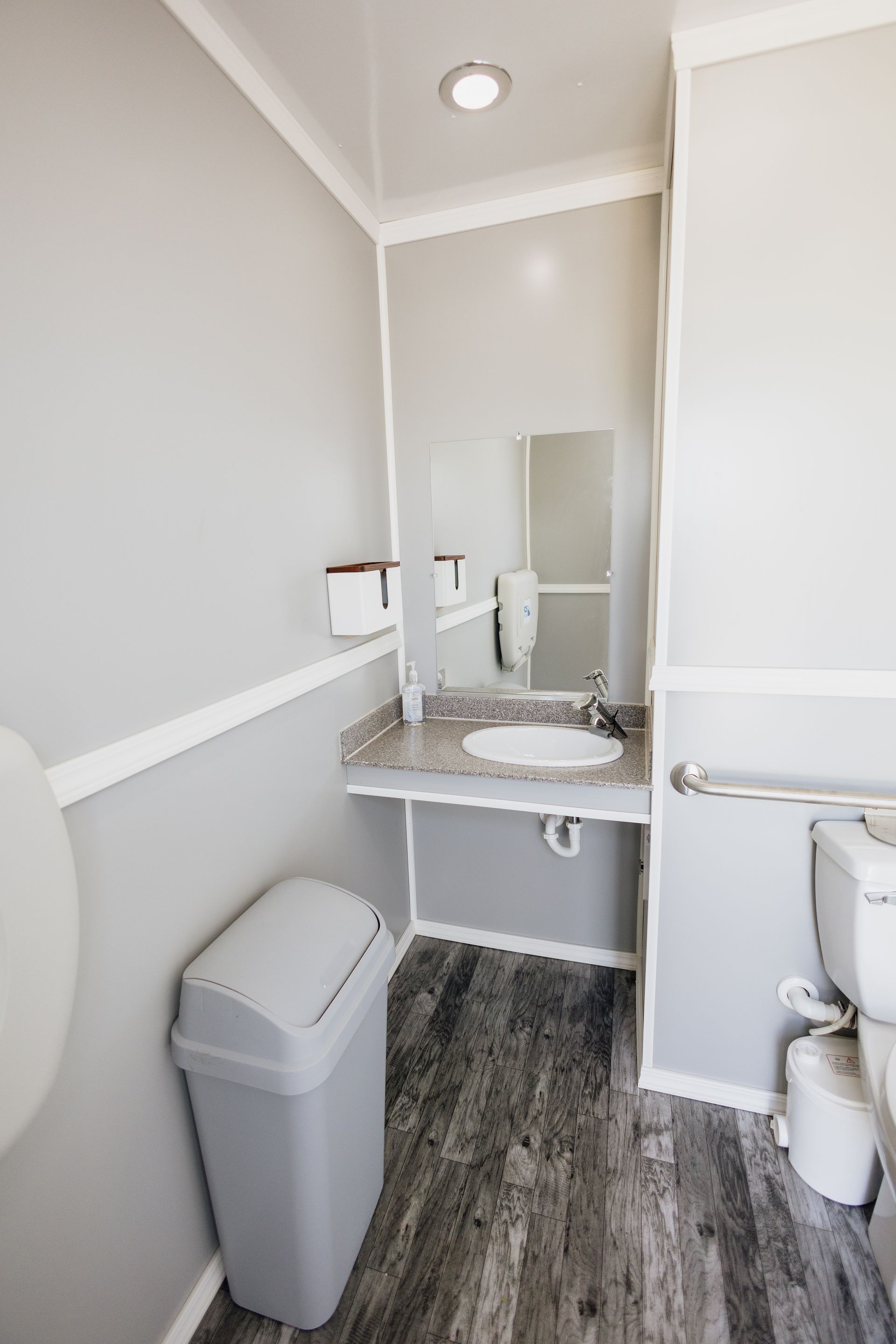 Small accessible restroom with a sink, mirror, toilet, gray walls, and wood-look flooring.