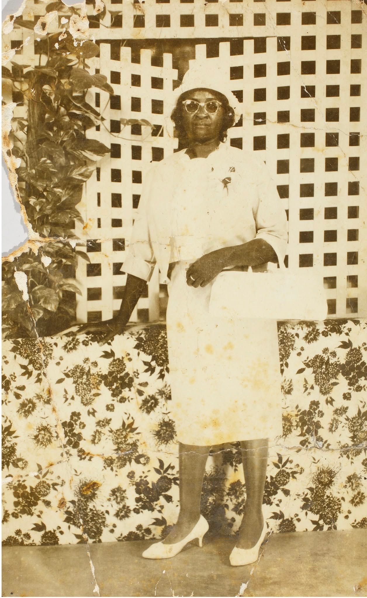 Person standing outdoors in a light dress and heels in front of a lattice wall and flowering bushes