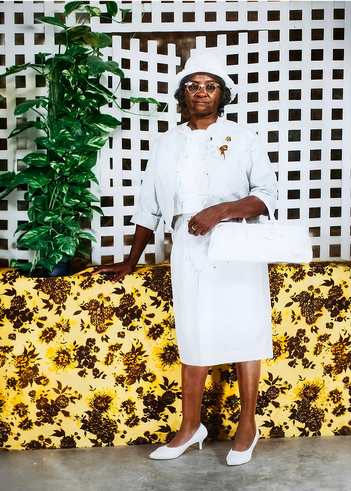 Person in white dress and heels standing indoors beside a plant, in front of a patterned wall and floral tablecloth