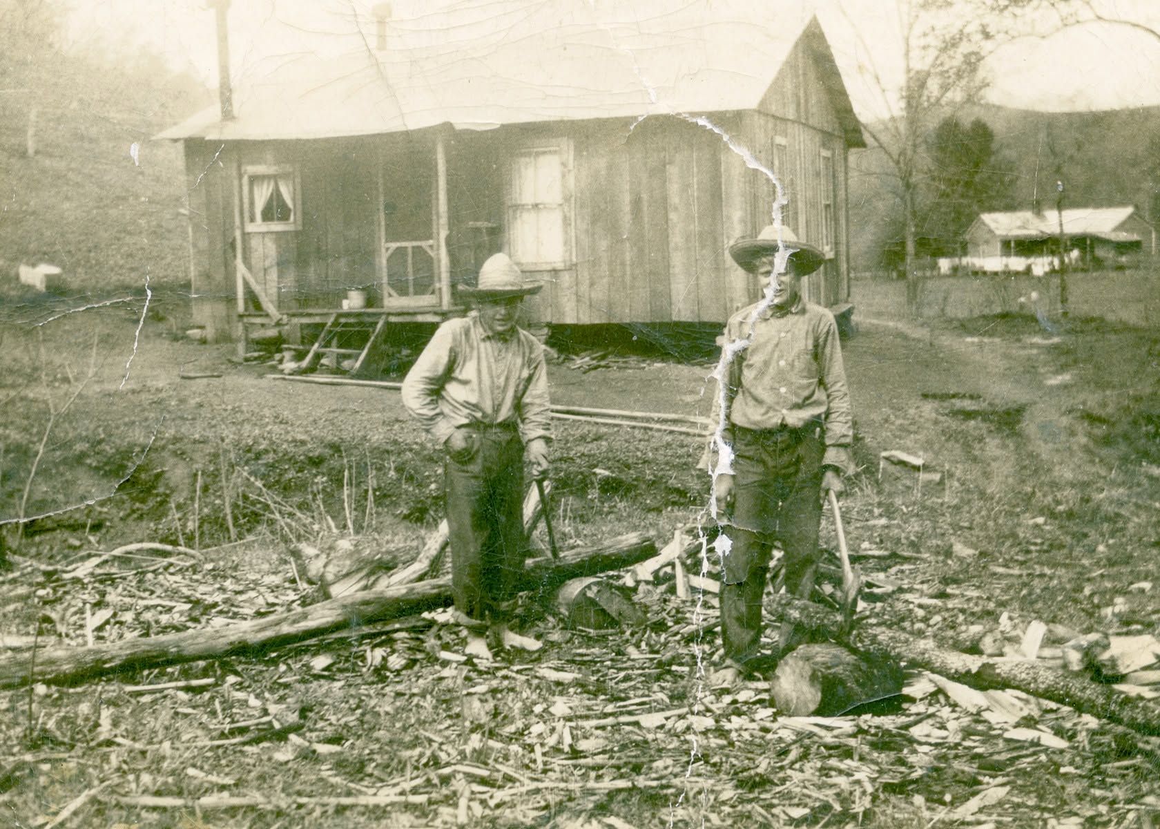 Two children stand in front of a rustic wooden cabin in a rocky, grassy yard.