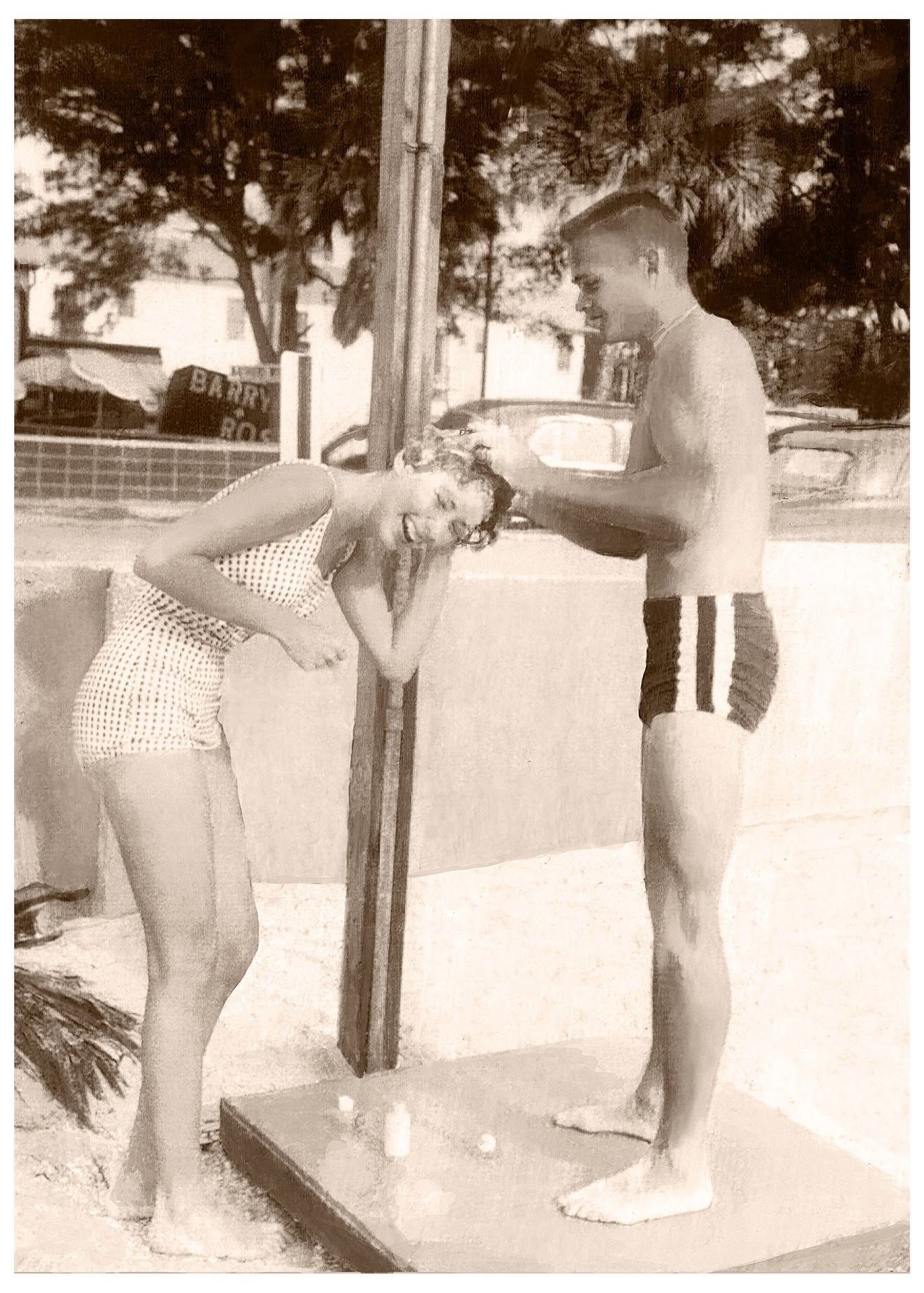 Two people at an outdoor shower or rinse station, one standing while the other bends forward under water.