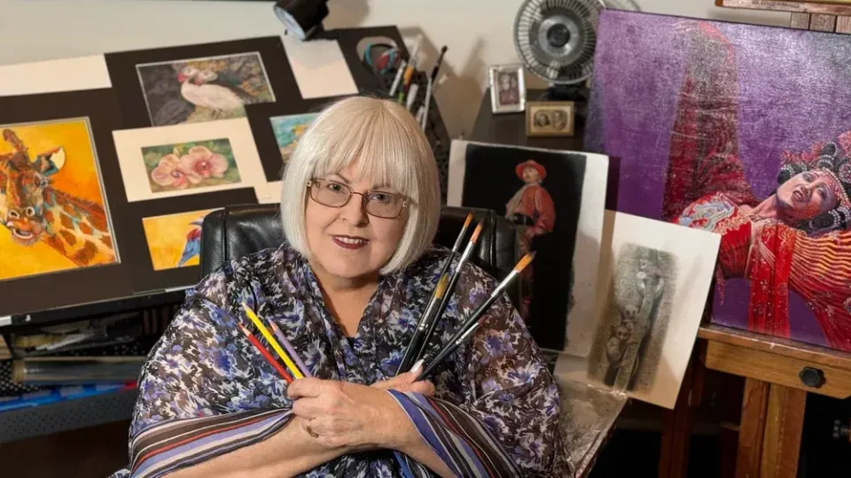 Artist seated in studio, holding paintbrushes and smiling amid colorful paintings and canvases