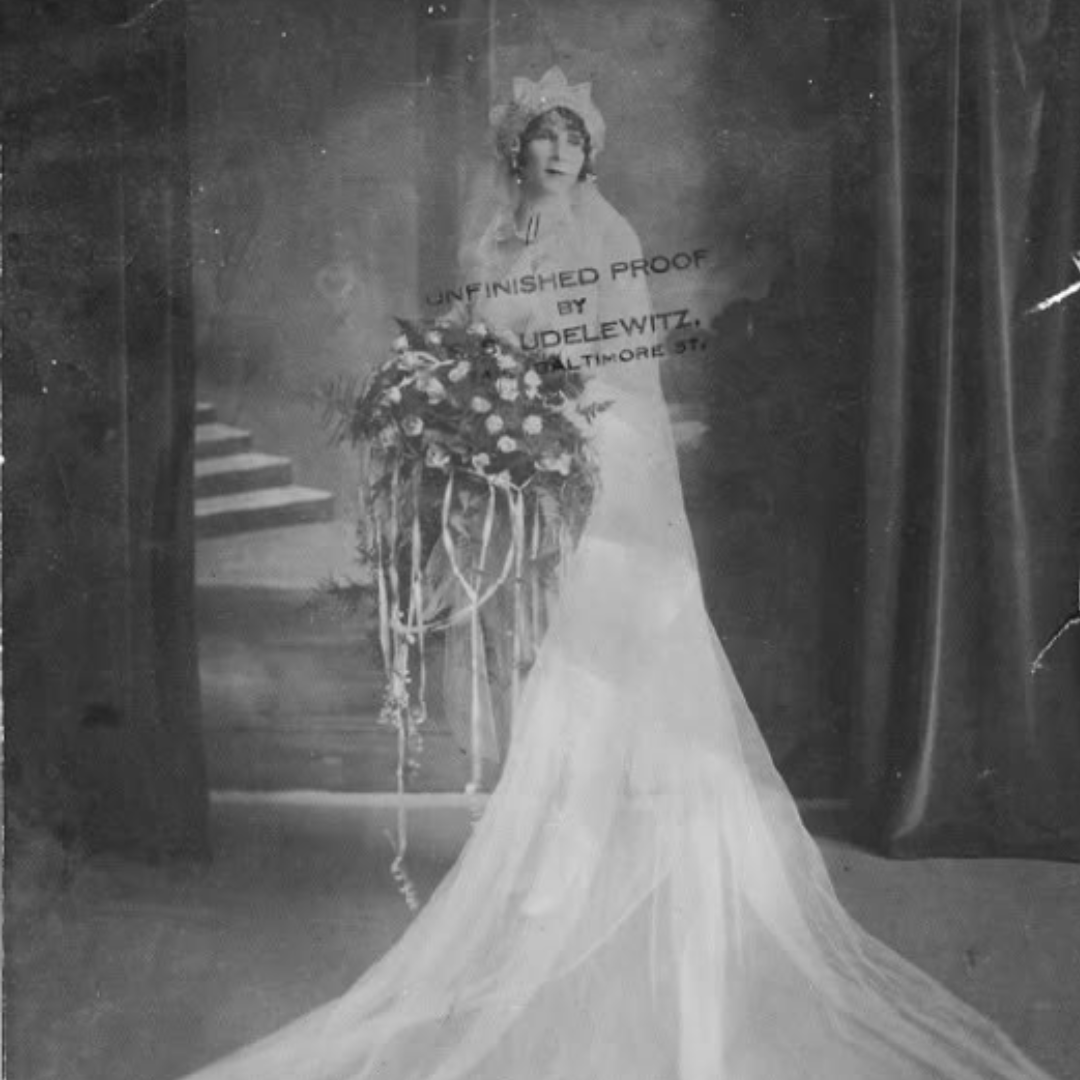 Person standing outdoors in a light dress and hat beside a lattice wall and flowering shrubs.