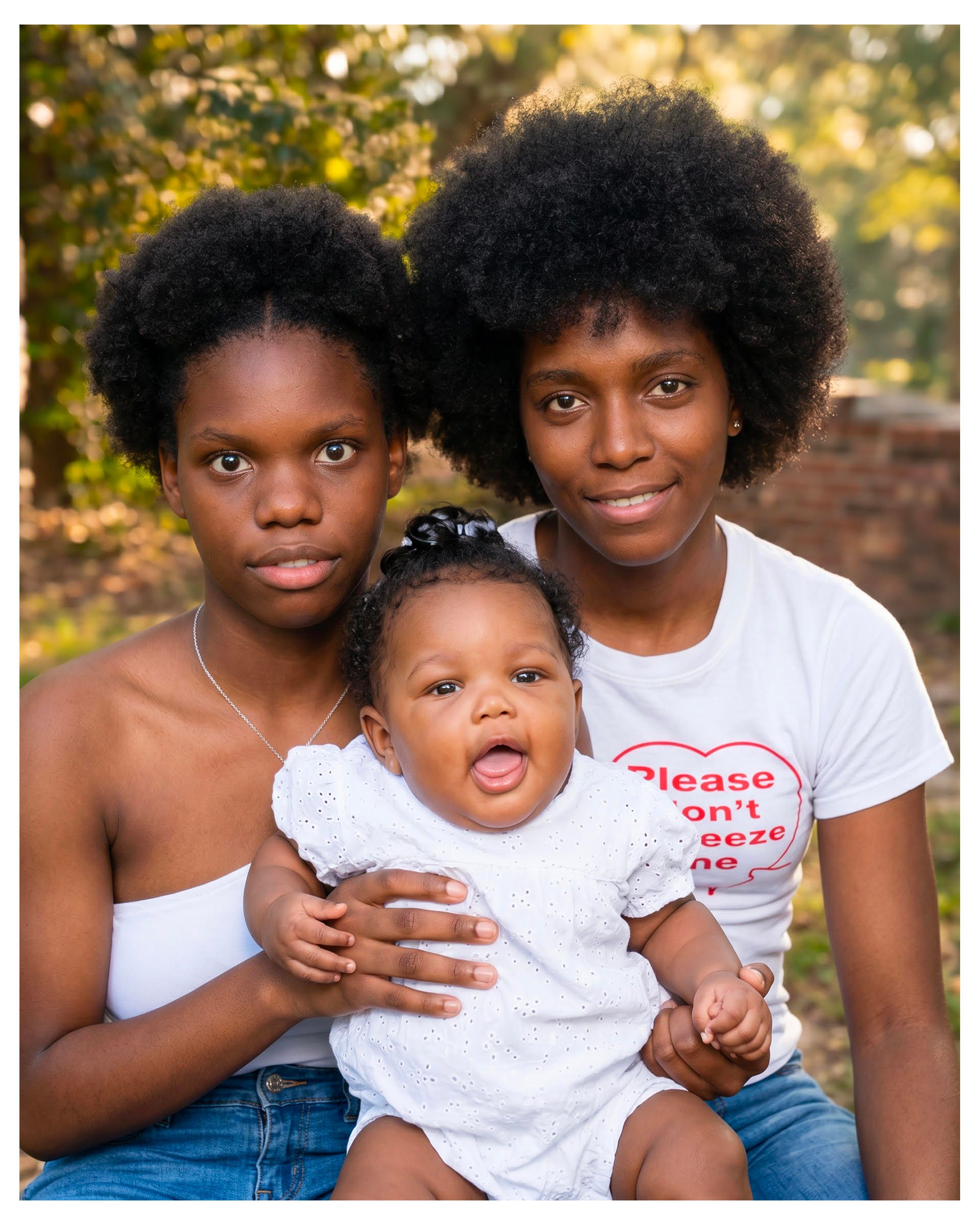 Two women sit with a baby outdoors, one holding the child, with a leafy background.