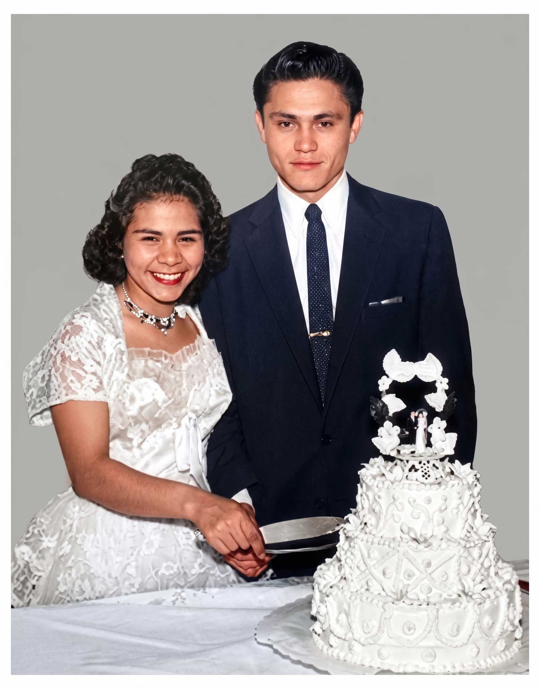 Couple in formal attire smiling beside a white wedding cake on a table