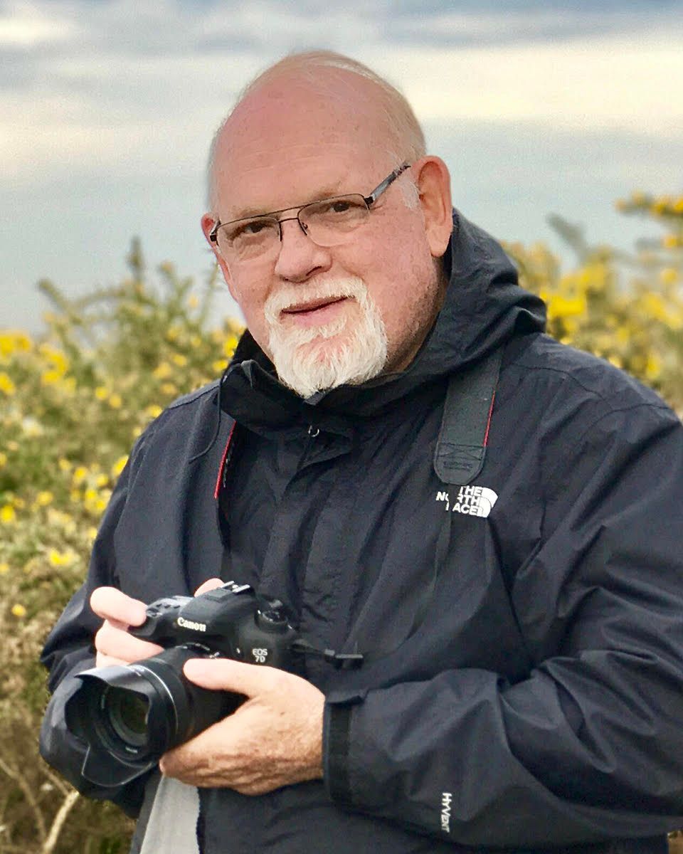 Smiling man in a black jacket holding a camera outdoors with yellow flowers in the background
