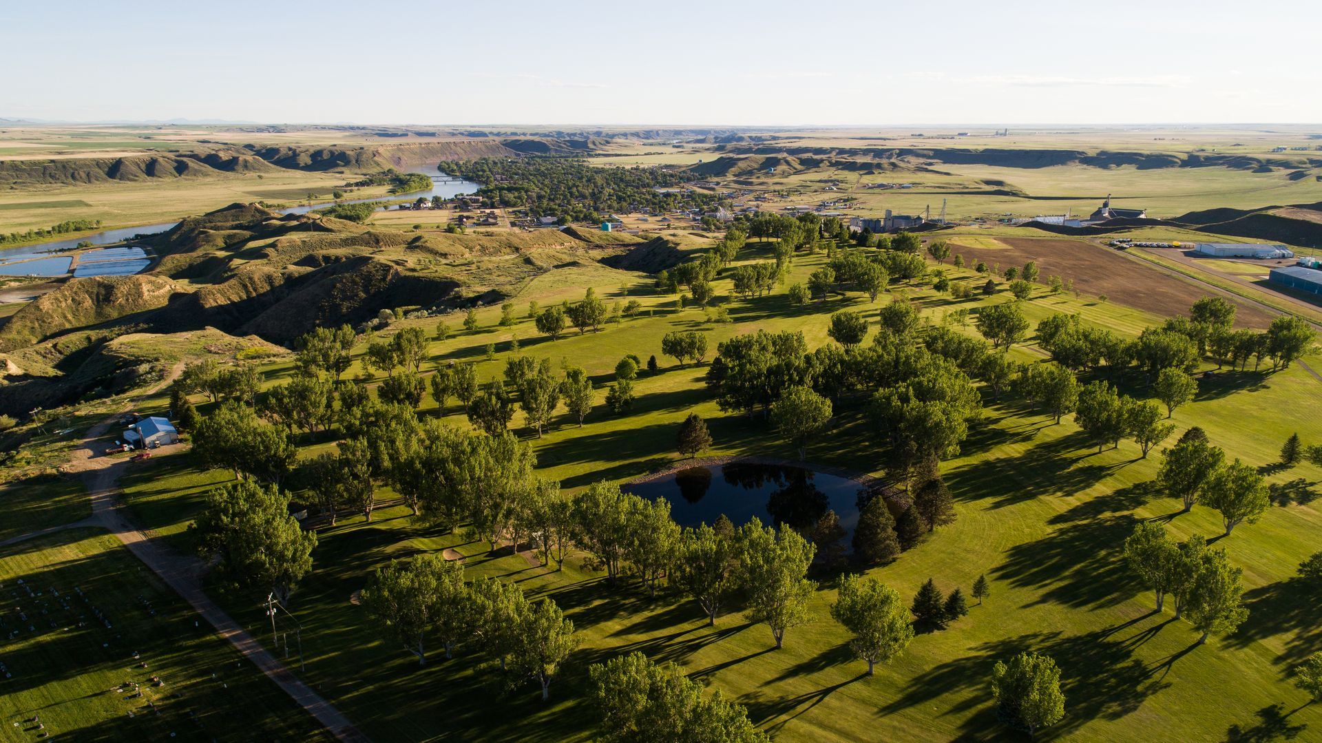 Aerial view of a sprawling park with lush trees, grassy hills, a small pond, and a river flowing through a distant valley.
