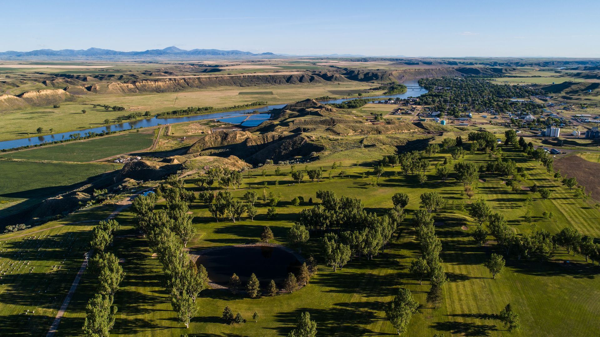 Aerial view of a river winding through a rural landscape with a park, scattered trees, and distant hills under blue sky.