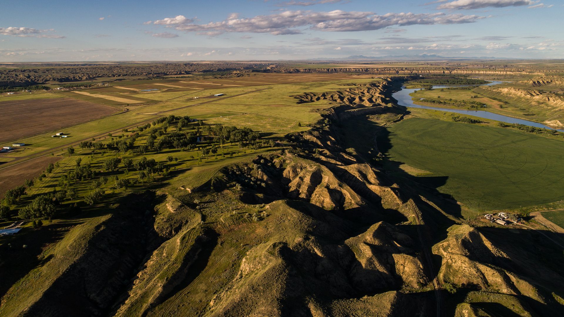 Aerial view of a winding river dividing a rugged, shadowed ridgeline from flat green fields and farmland under a blue sky.