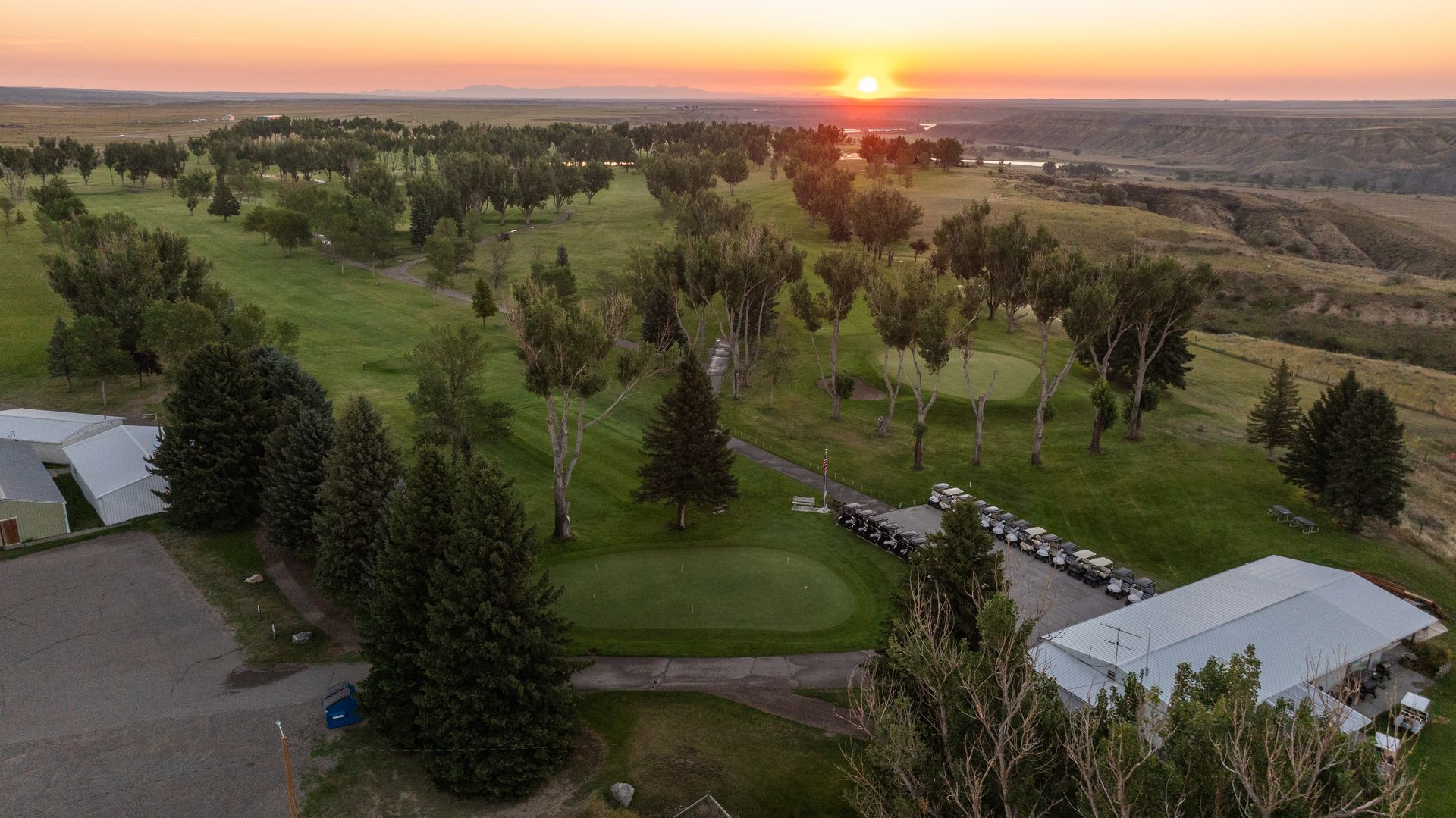 Aerial view of a golf course at sunset, with a clubhouse, golf carts, and trees lining the green fairways.