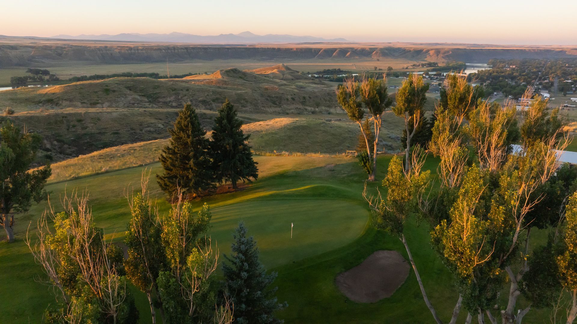 A scenic high-angle view of a sunlit golf green with a sand trap, surrounded by trees and hilly, rolling terrain at sunset.
