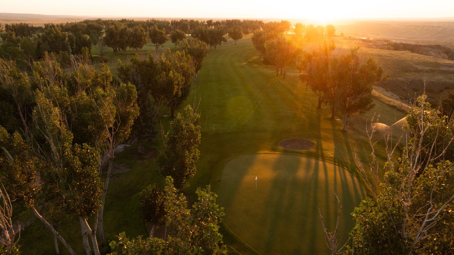 An elevated view of a golf course fairway lined with trees at sunset, with a green visible in the foreground.