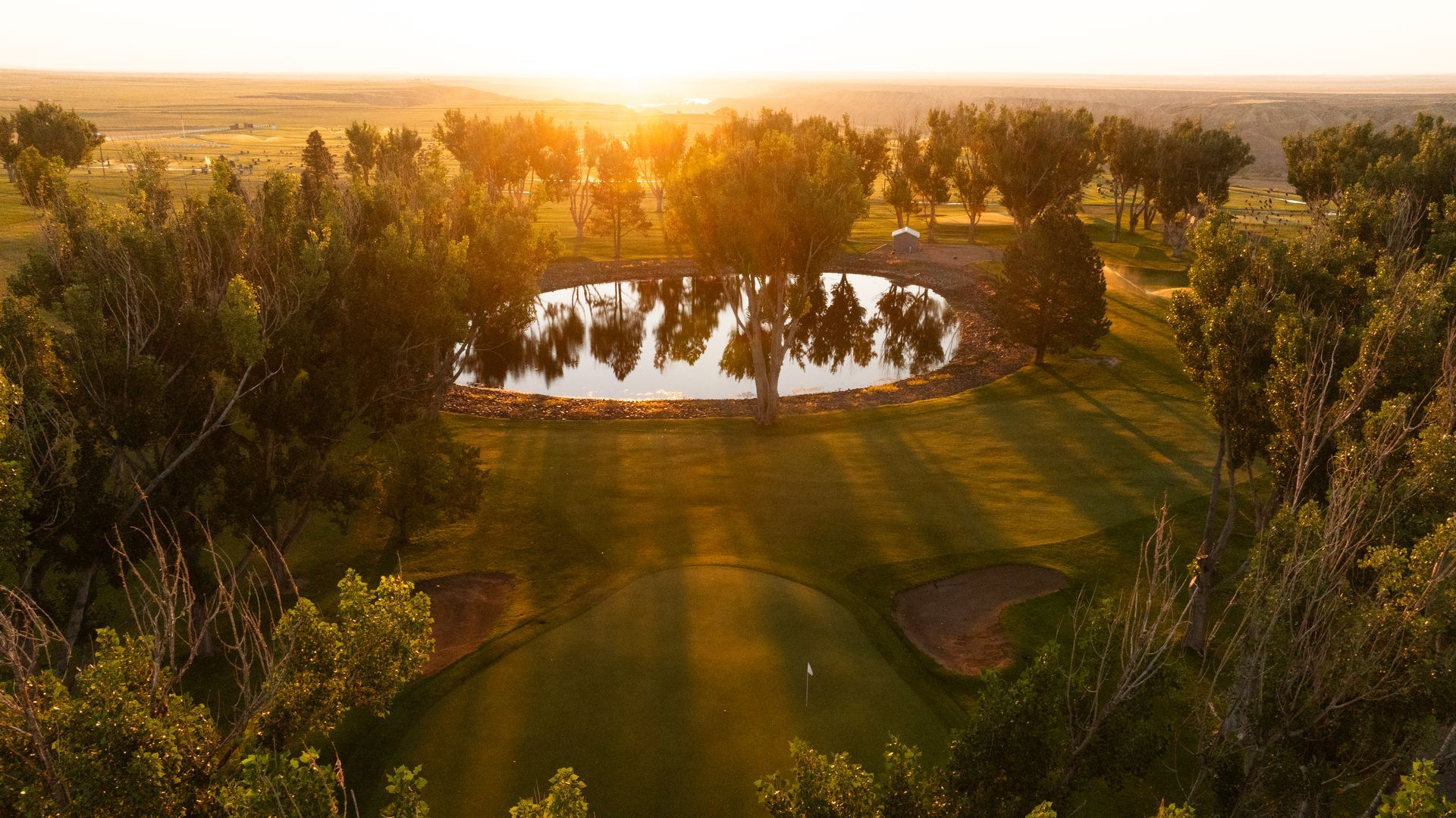 A golf course green sits before a pond, framed by trees under a bright, golden sunset.