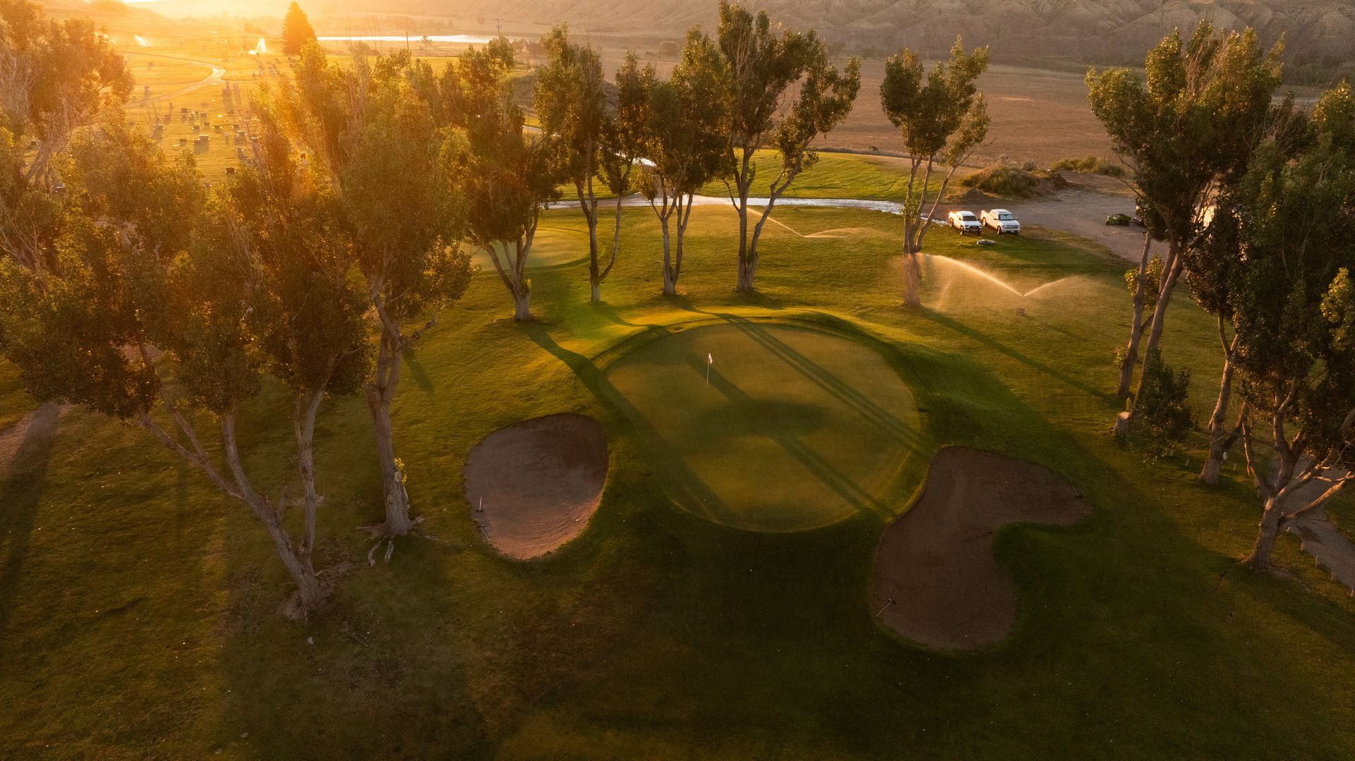 An elevated view of a golf green surrounded by sand bunkers and trees, illuminated by warm golden hour sunlight.