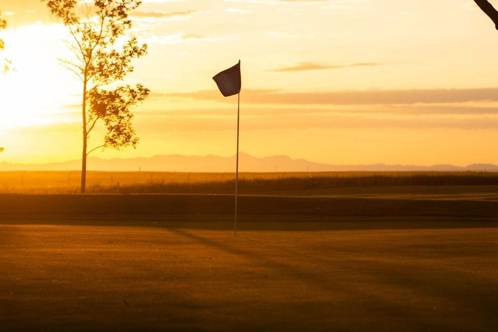 A golf flag stands on a putting green at sunset, with distant mountains silhouetted against a golden sky.