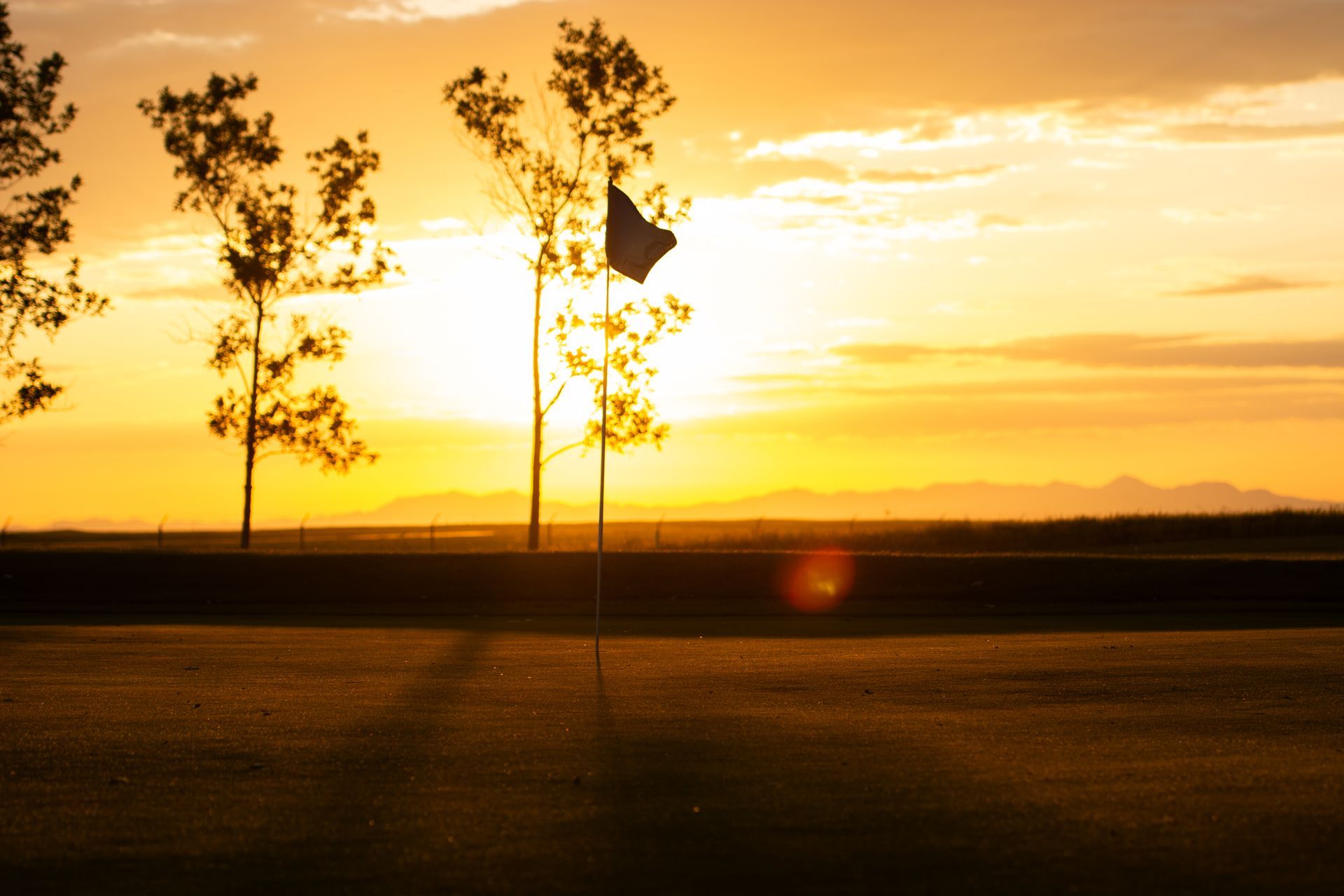 A silhouette of a golf flag on a course against a vibrant, golden sunset with distant mountains.