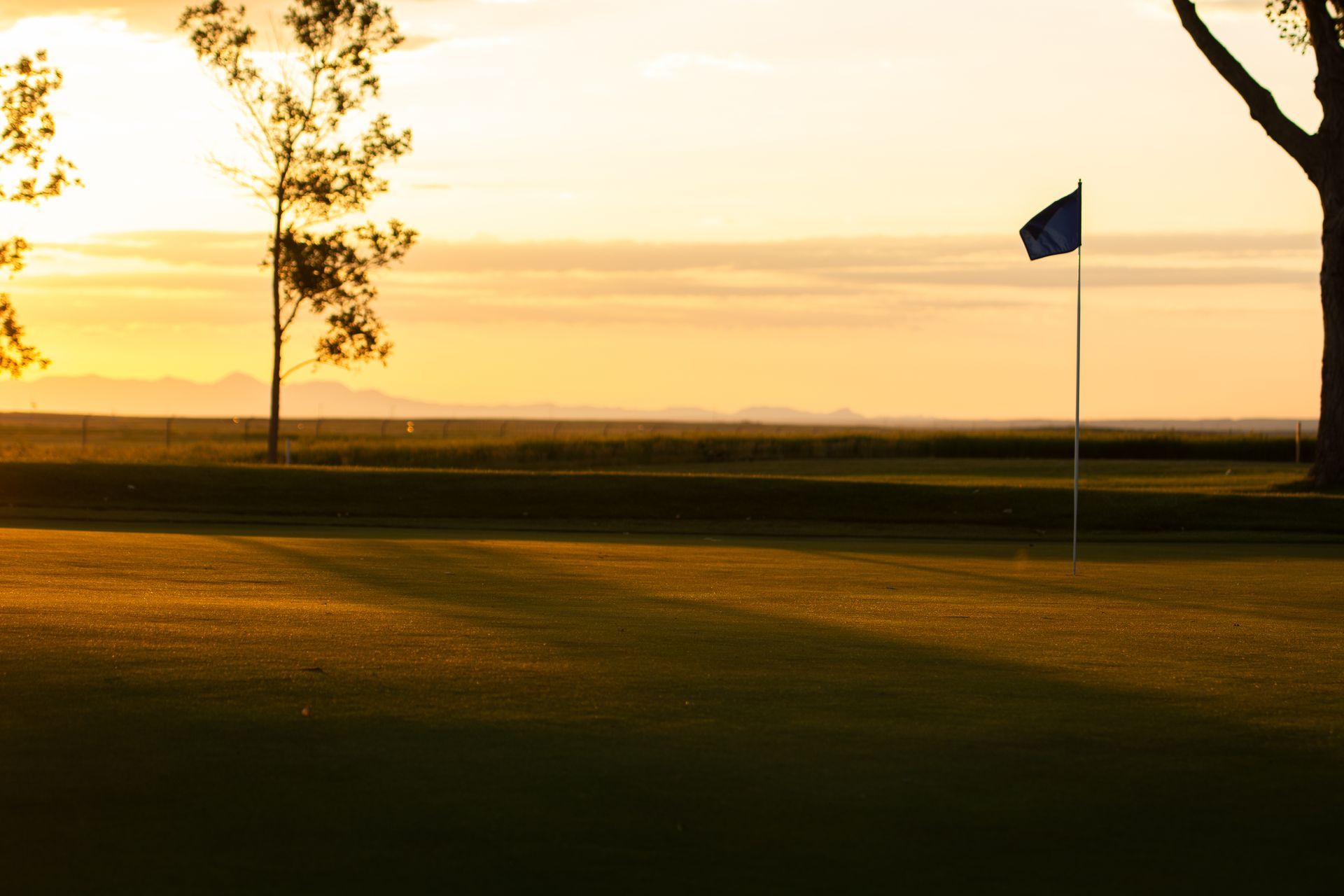 A golf course green at sunset with a blue flag on a pole, silhouetted trees, and mountains in the distance.