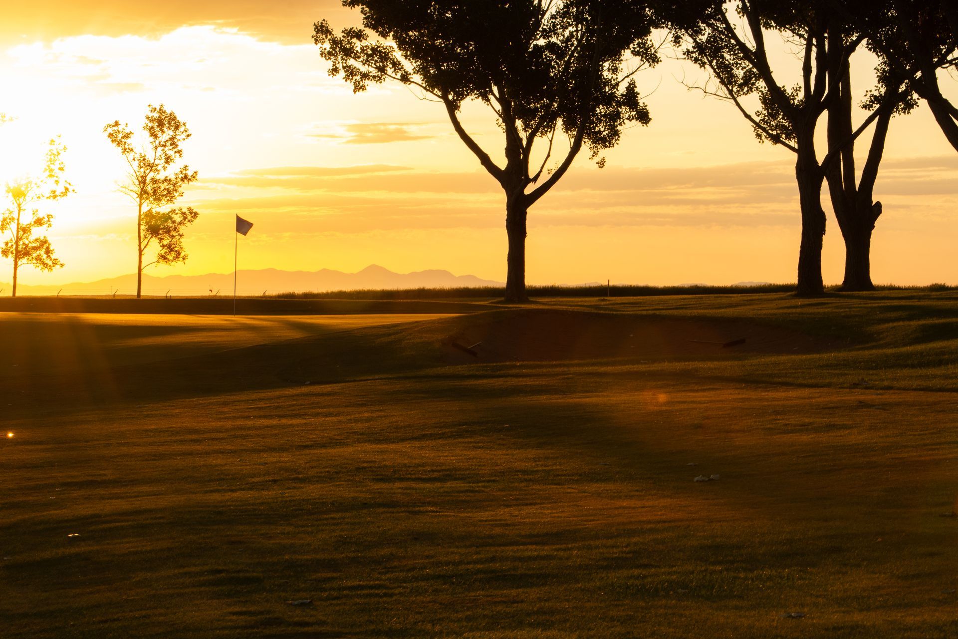 A golf course green with a flag pole at sunset, silhouetted by trees against a golden horizon.