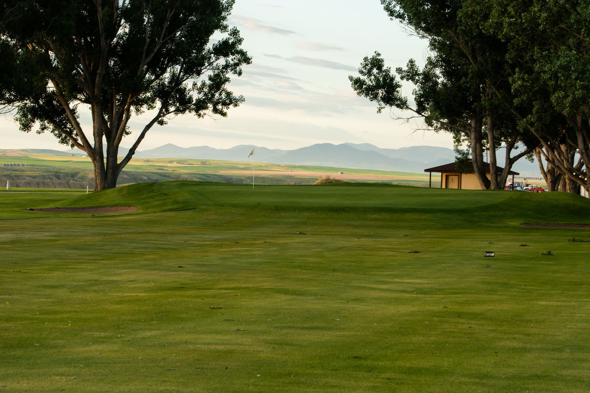 A green golf course fairway leads toward a putting green, framed by trees on both sides with distant mountains at sunset.