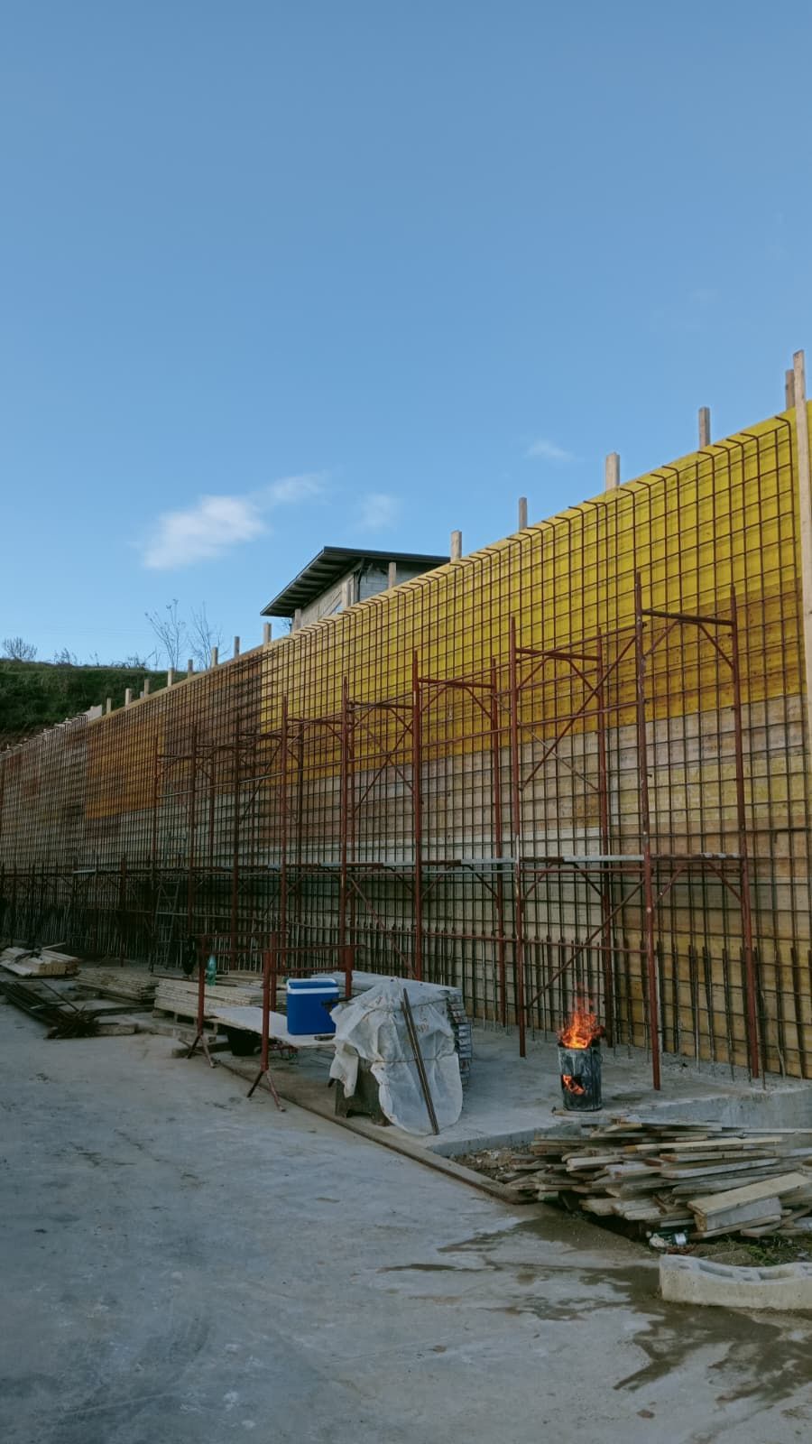 Lungo muro di contenimento in cemento in costruzione, con impalcature a rete gialla e attrezzi, lungo una strada sterrata.