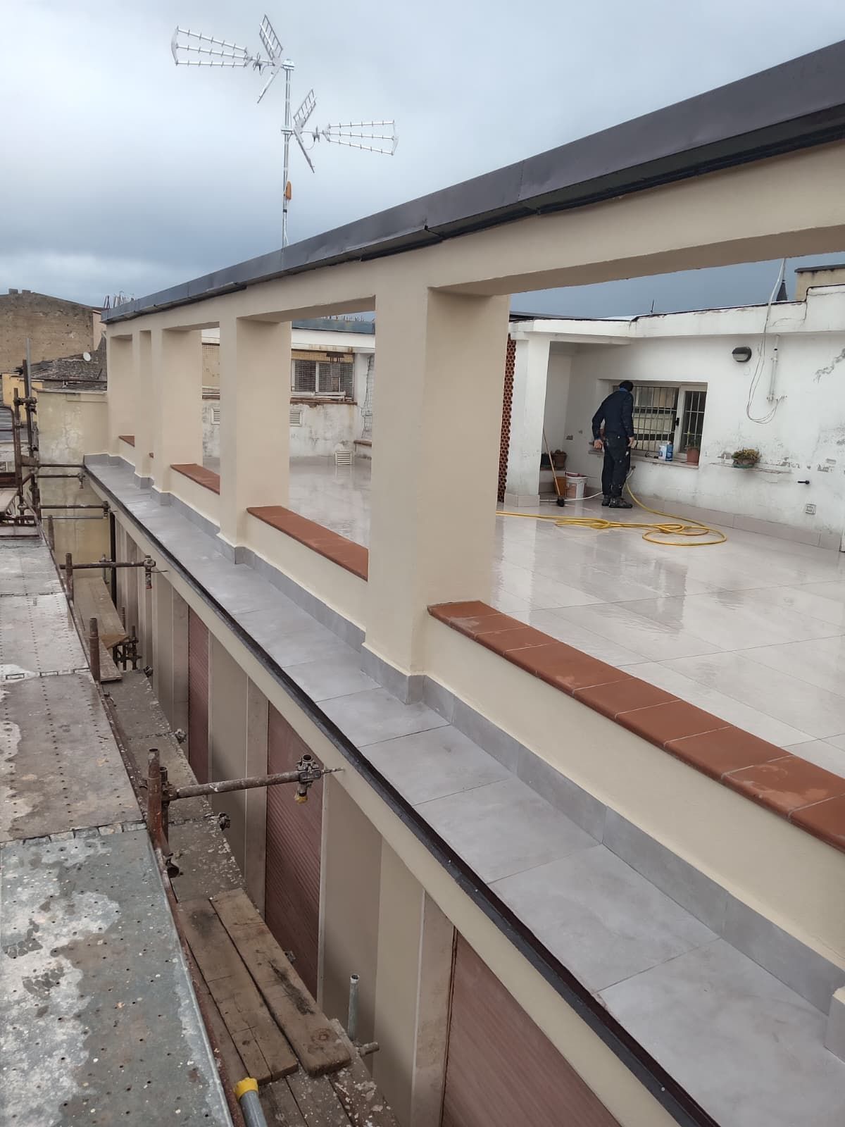 Terrazza sul tetto con pareti color crema, pavimento grigio e una persona che lavora vicino a un edificio bianco sullo sfondo.