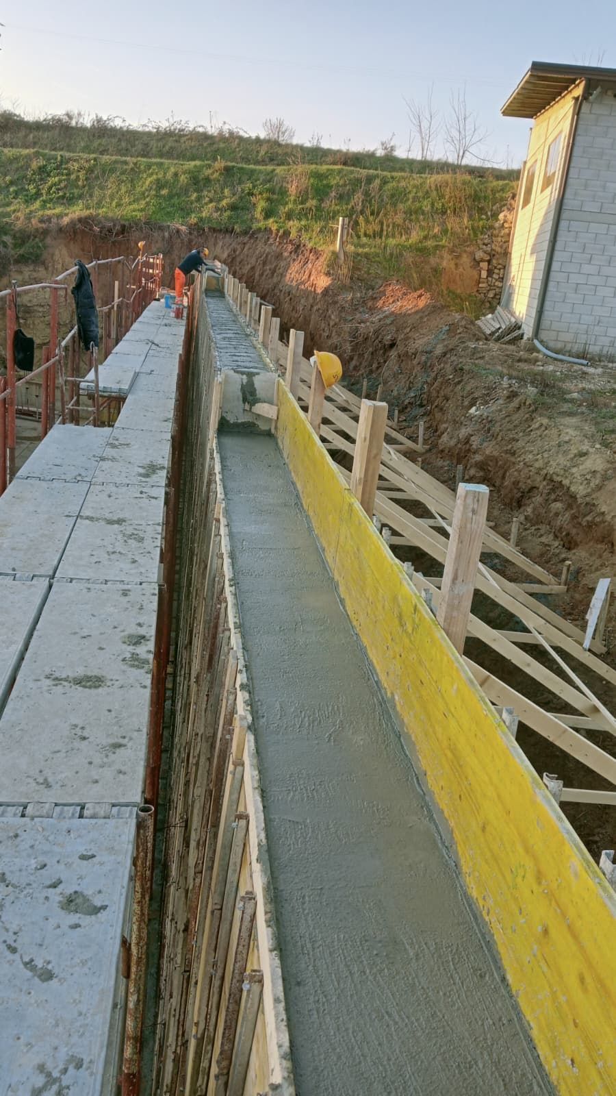 Rampa esterna in cemento con ringhiere di sicurezza gialle, adiacente a un muro di mattoni e a un pendio.