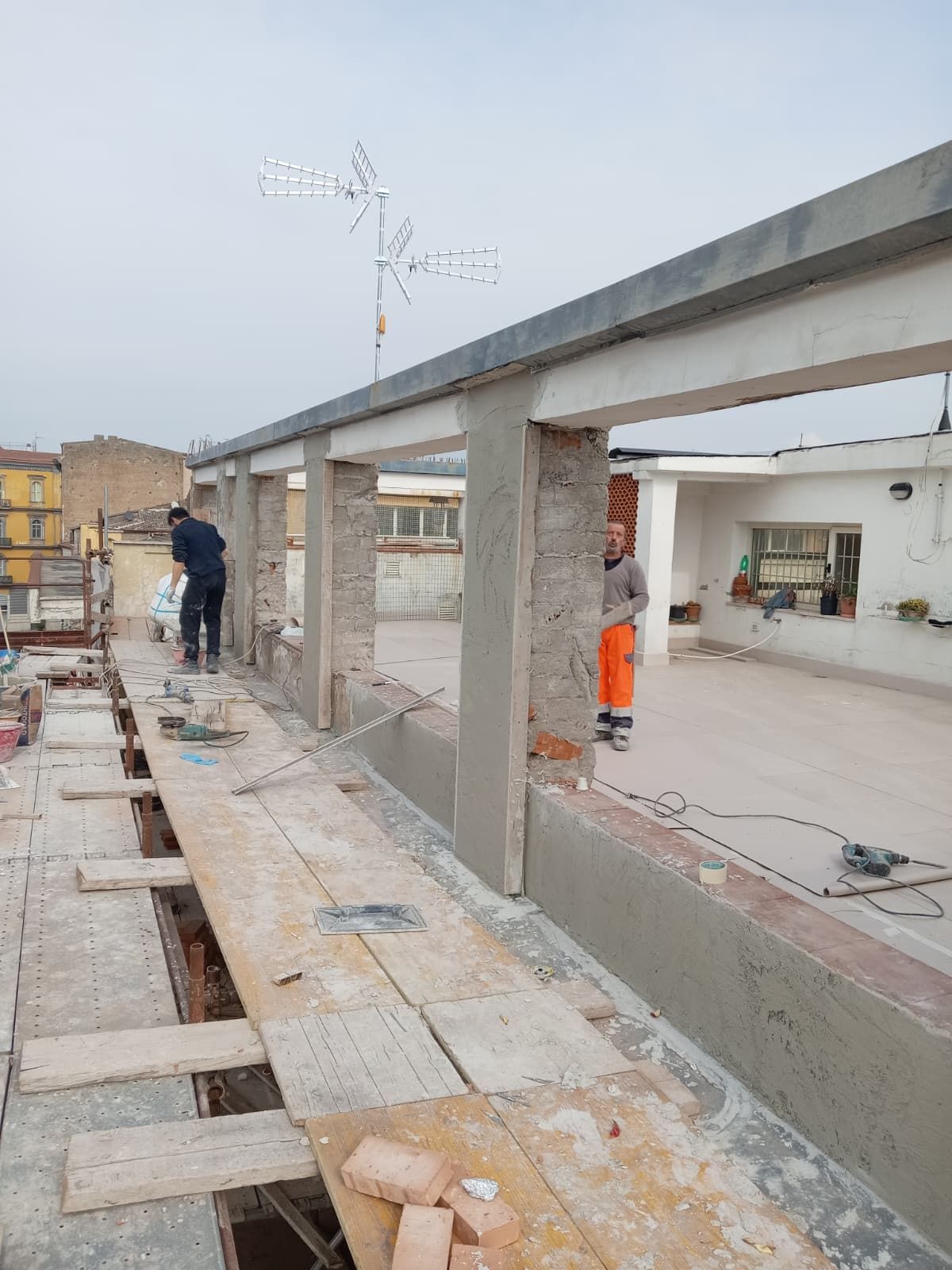 Passerella del cantiere accanto alla struttura in cemento armato di un edificio, con operai e assi di legno lungo il bordo.