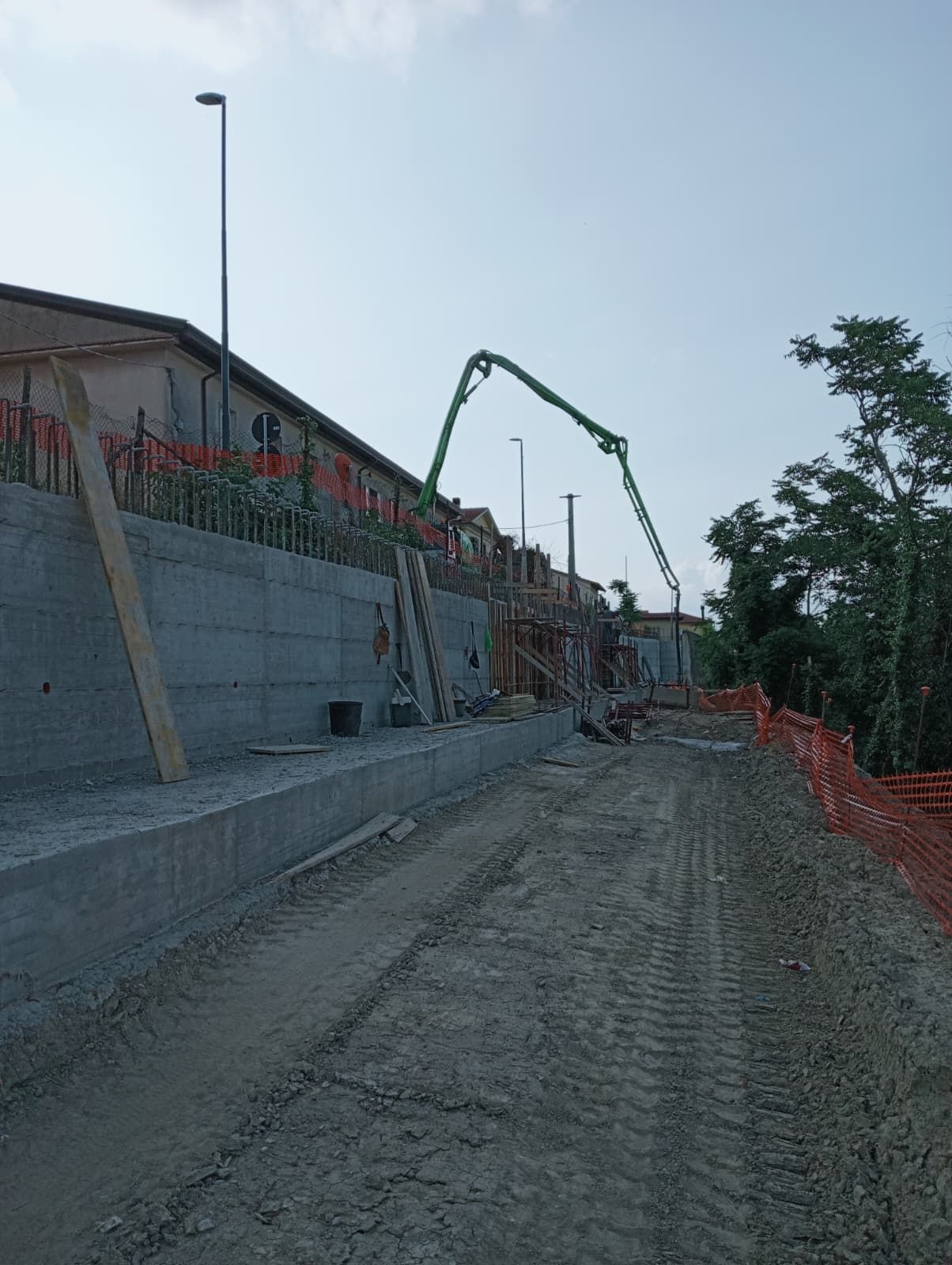 Cantiere edile con muro di cemento, strada sterrata, recinzione arancione e un'autobetoniera sullo sfondo.