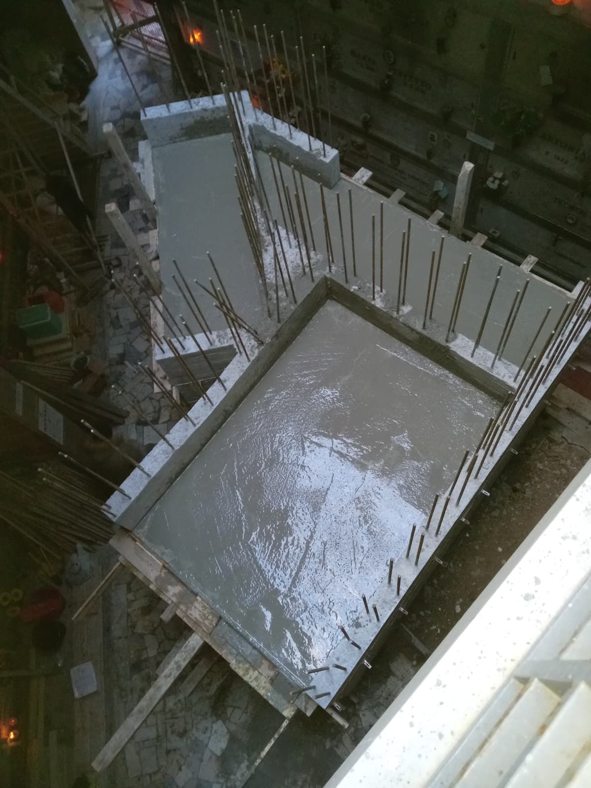 Cantiere edile con soletta in cemento fresco, barre d'armatura sporgenti e casseratura attorno allo scavo di fondazione.