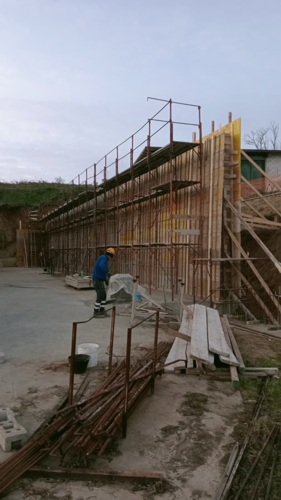 Cantiere edile con un lungo muro di cemento, impalcature in tondini di ferro e un operaio in divisa blu vicino a materiali accatastati.