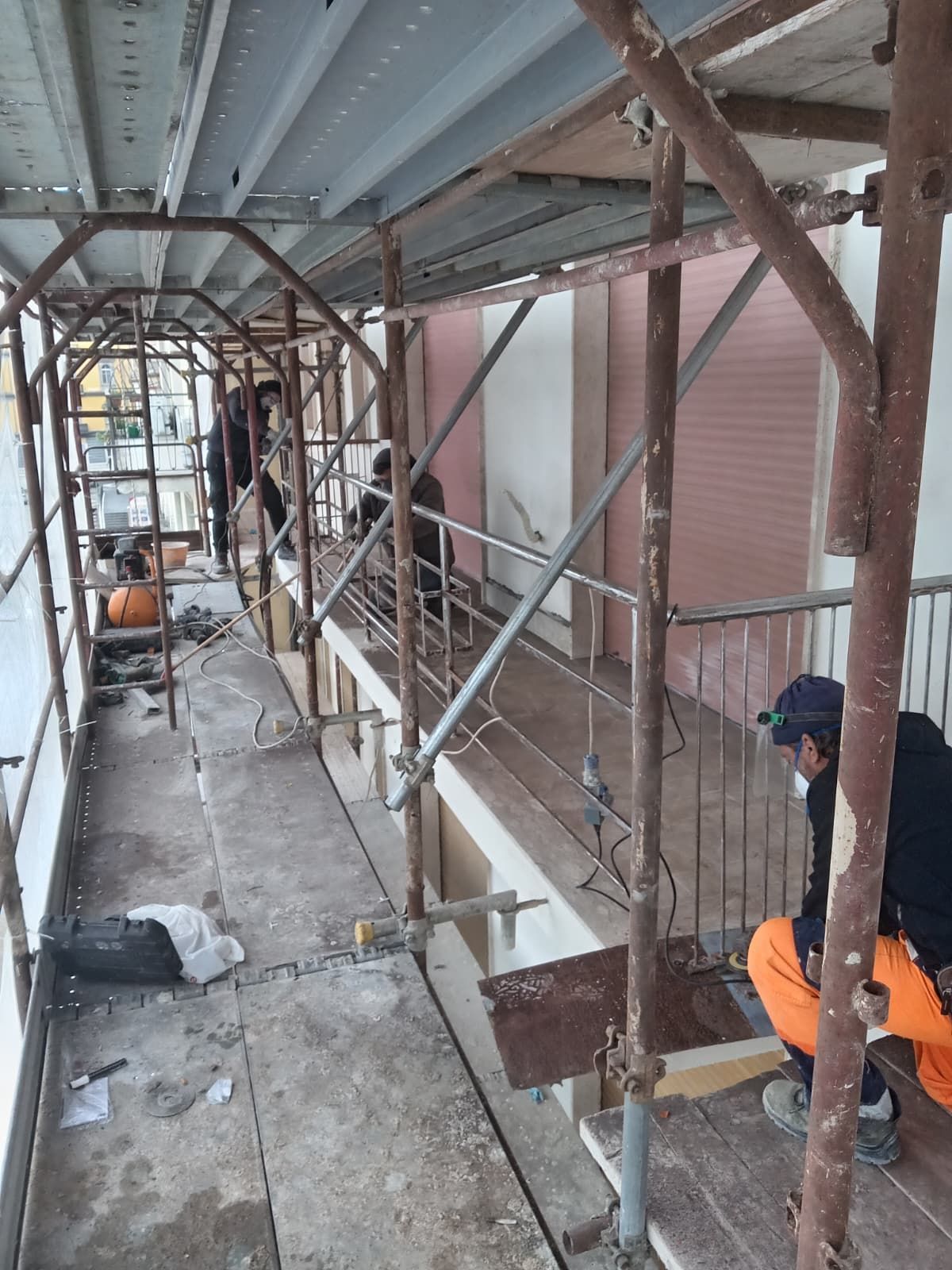 Passerella fiancheggiata da impalcature accanto a un edificio, con due operai in tuta arancione intenti a riparare il muro.