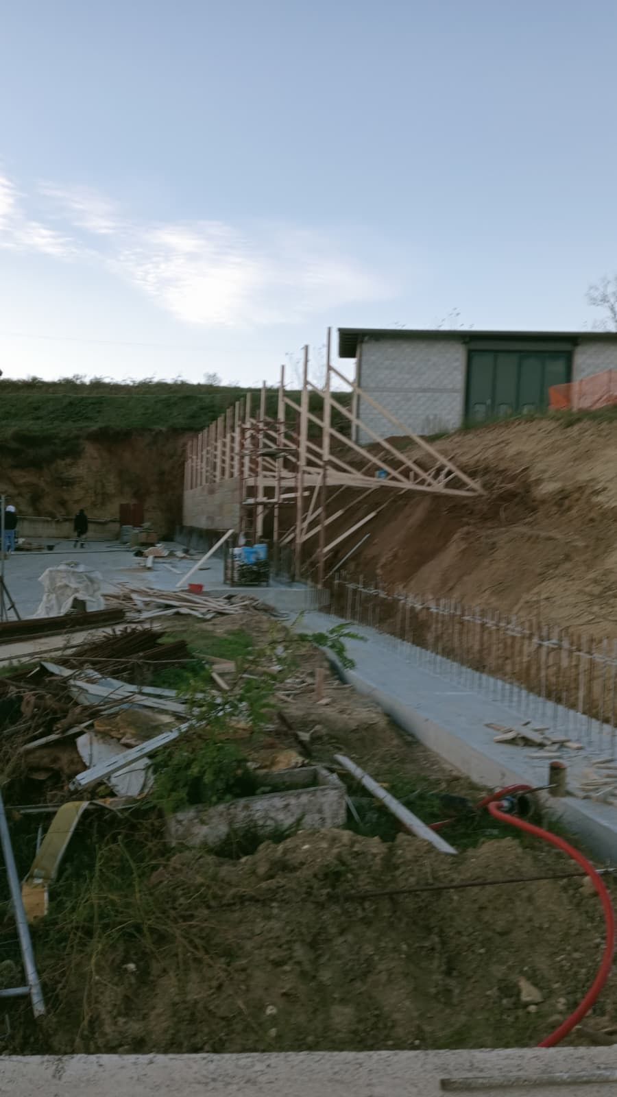 Cantiere edile all'aperto con impalcature di legno accanto a un edificio e uno scavo di terra in primo piano.